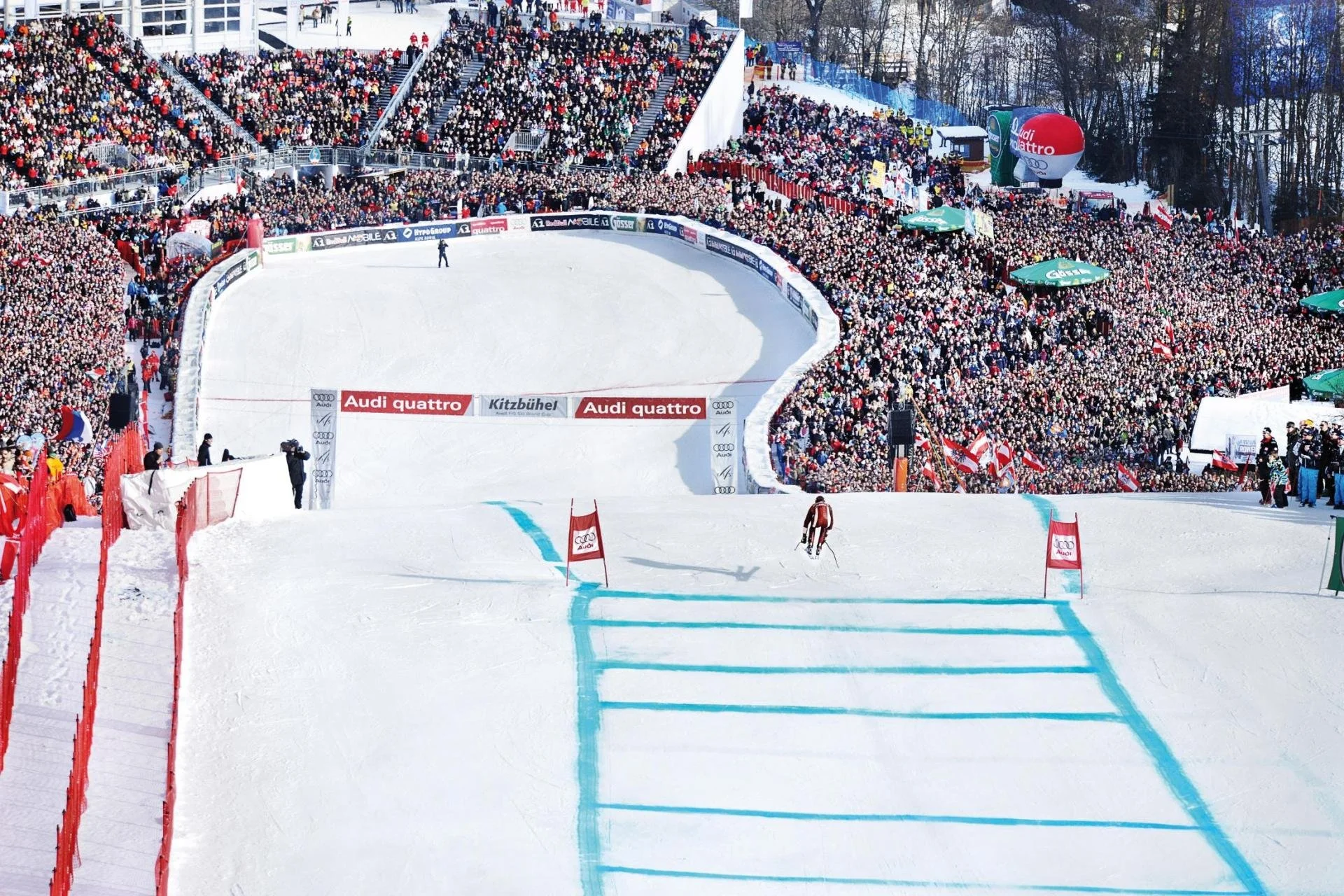 Skiers crossing the finish line of the Hahnenkamm downhill race on a snow-covered slope in Kitzbuhel, with a large crowd of spectators in the background.