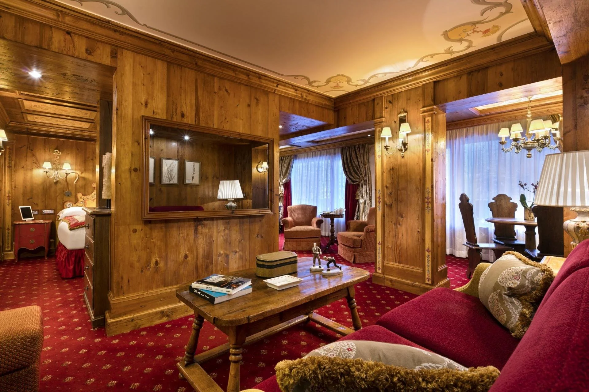 A cozy living room with wooden walls and ceiling, red patterned carpet, armchairs, a sofa with patterned cushions, a wooden coffee table with books and toys, a table lamp, and curtains covering large windows.