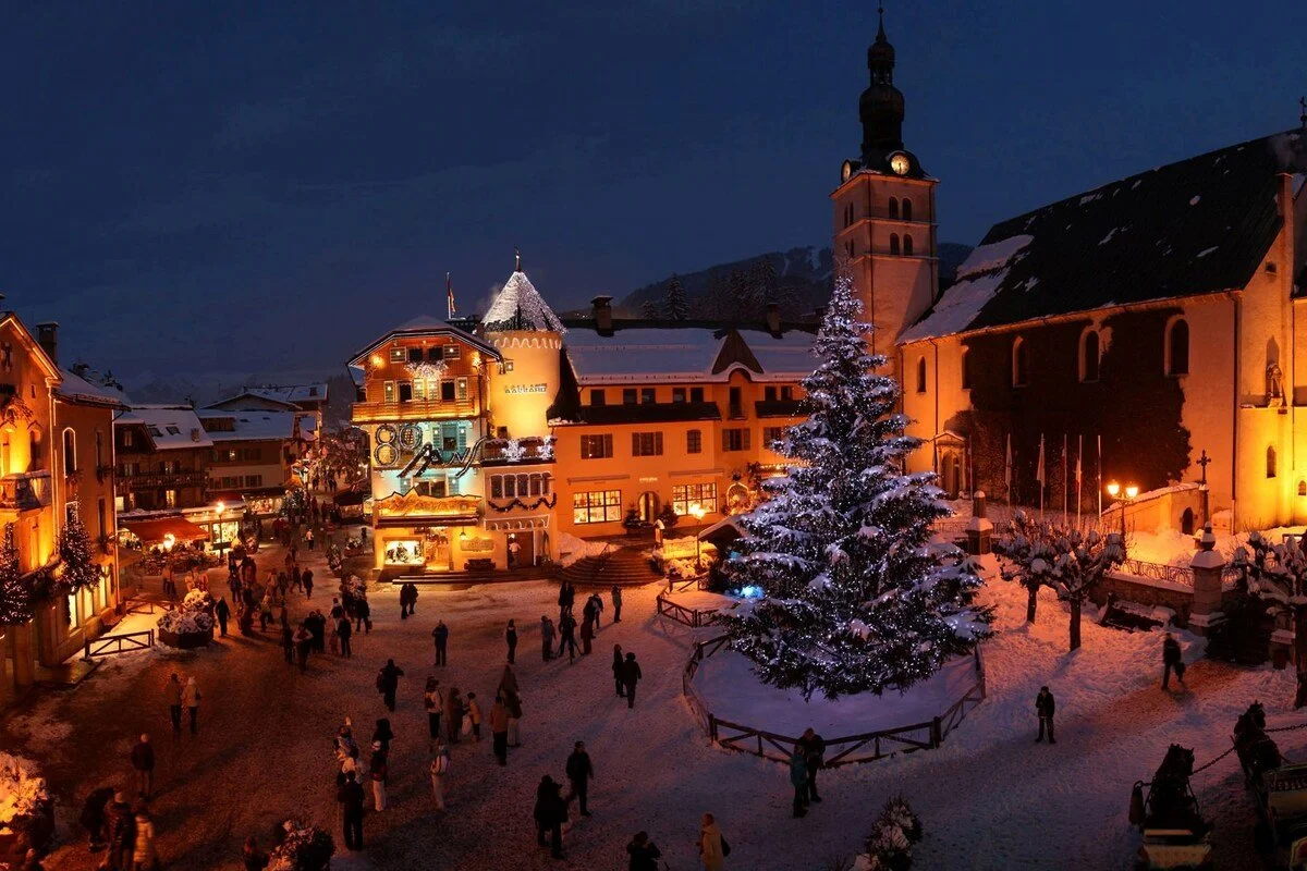 A snowy Megeve village with Christmas lights, a decorated tree, and a church tower at night.