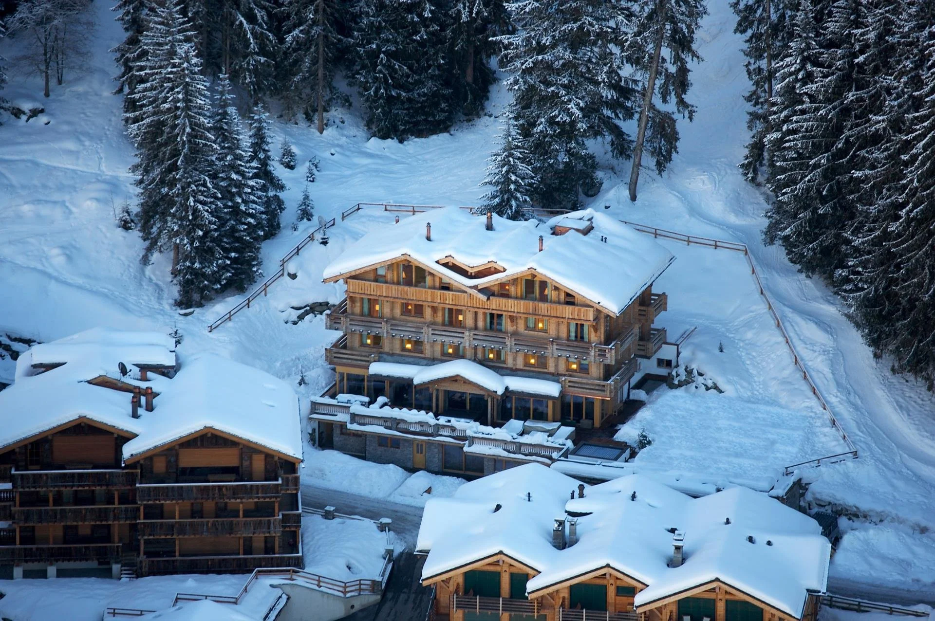 A snow-covered mountain landscape featuring several wooden chalets and a dense forest of pine trees.