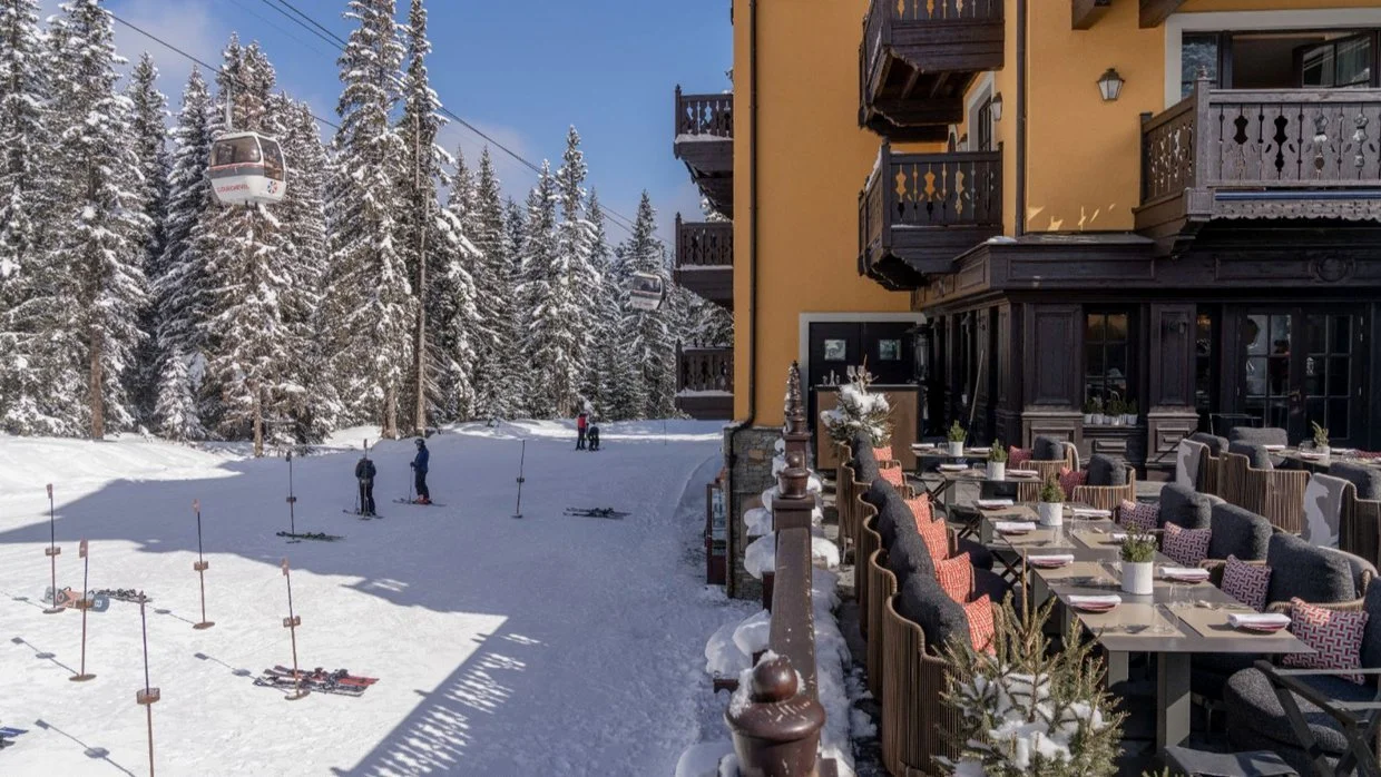 A snowy outdoor area with skiers and snowboards, tall snow-covered pine trees, ski lifts, and a patio with tables and chairs outside a yellow building.