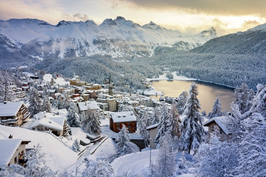 Snow-covered St Moritz village in a mountainous region during winter with St Moritz lake and dense forest in the background.