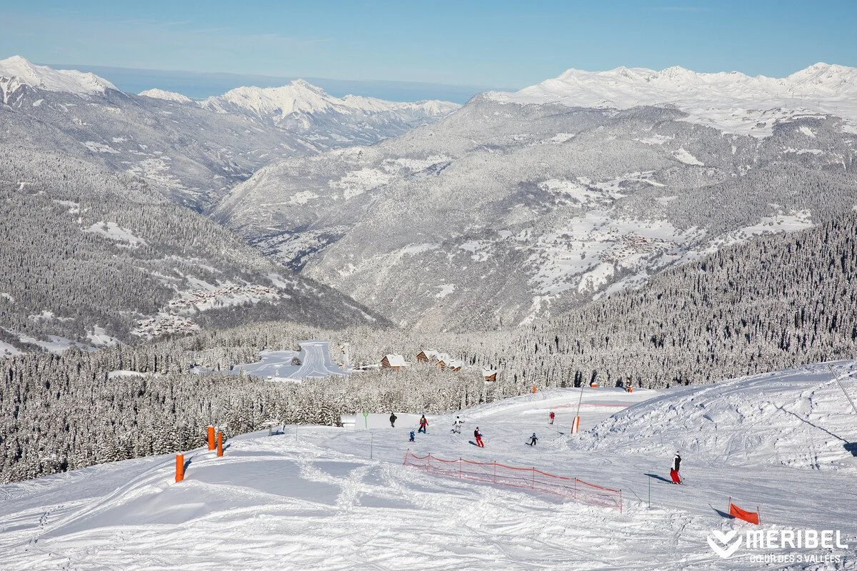 Skiers on a snowy mountain slope in Meribel with a forest and mountain range in the background.