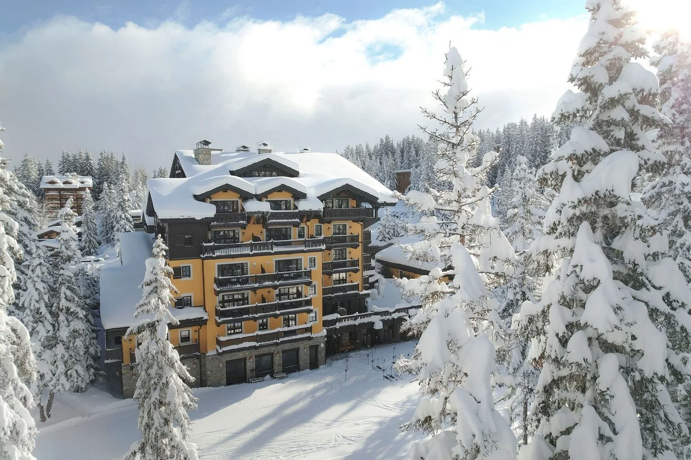 Snow-covered mountainside hotel with yellow and dark wooden exterior, surrounded by snow-laden trees under a cloudy sky.