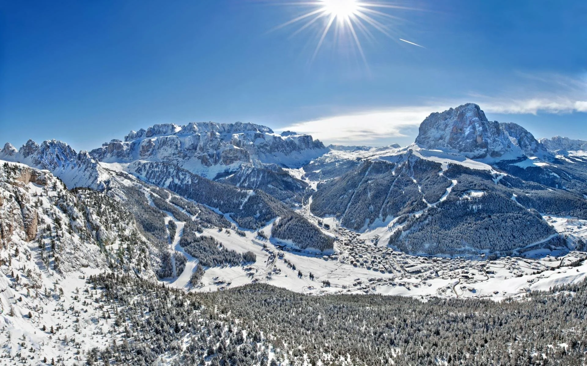 Snow-covered mountains and a village in a valley under a bright sun in a clear blue sky.