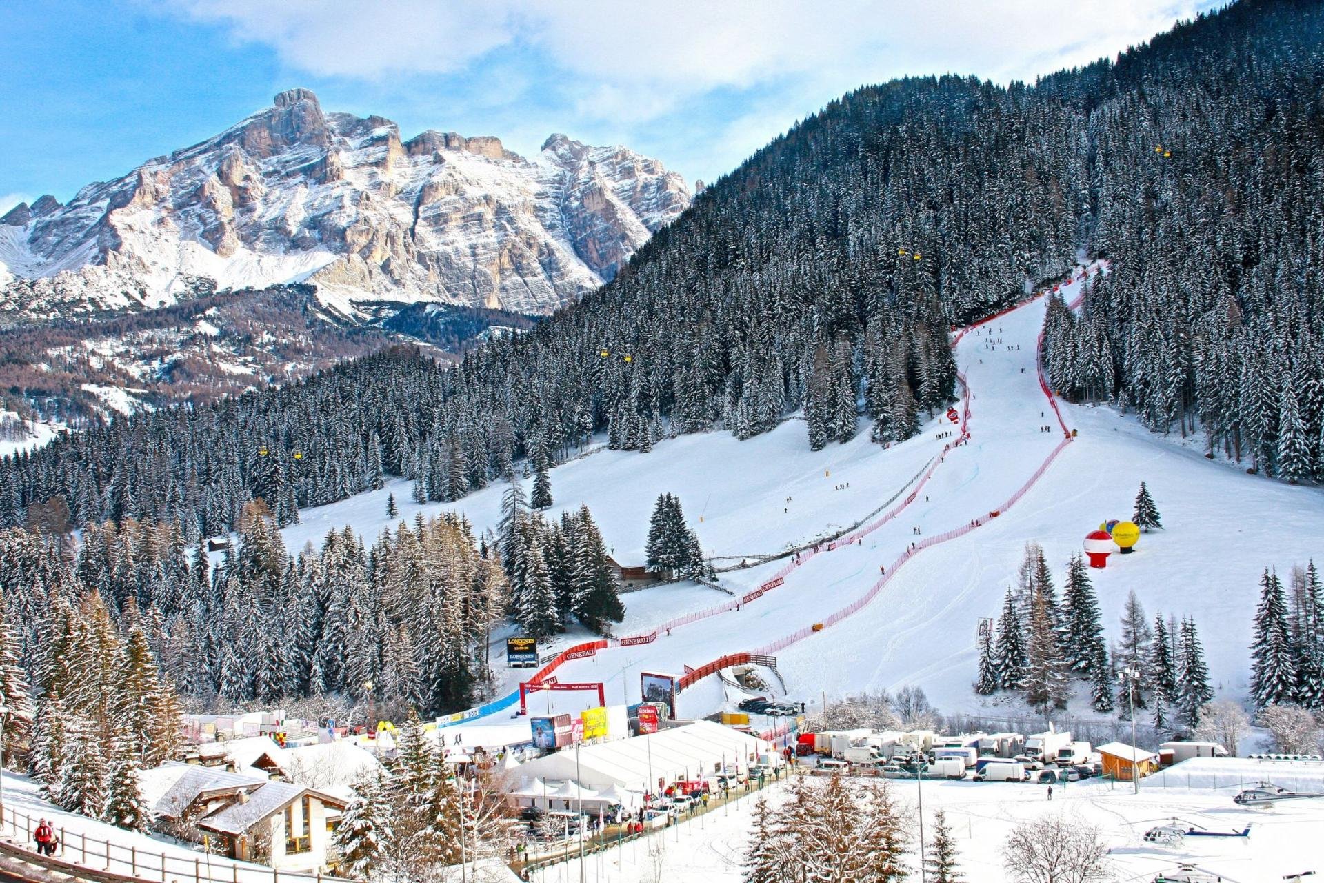 Snow-covered ski slope with skiers, surrounded by dense pine trees and mountains in the background, with a parking area and buildings at the bottom.