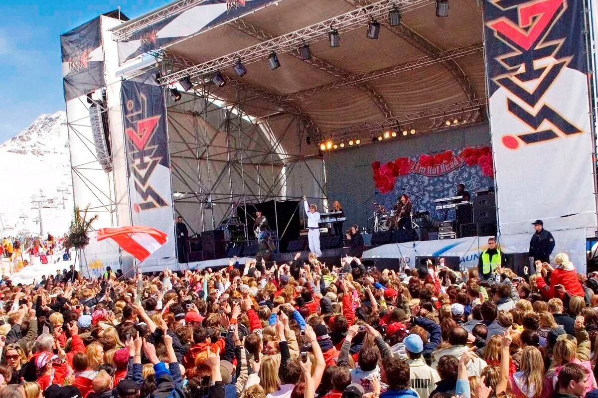 A large outdoor concert stage in a snowy Ischgl, with a band performing and a crowd of people watching and taking photos, some waving flags.