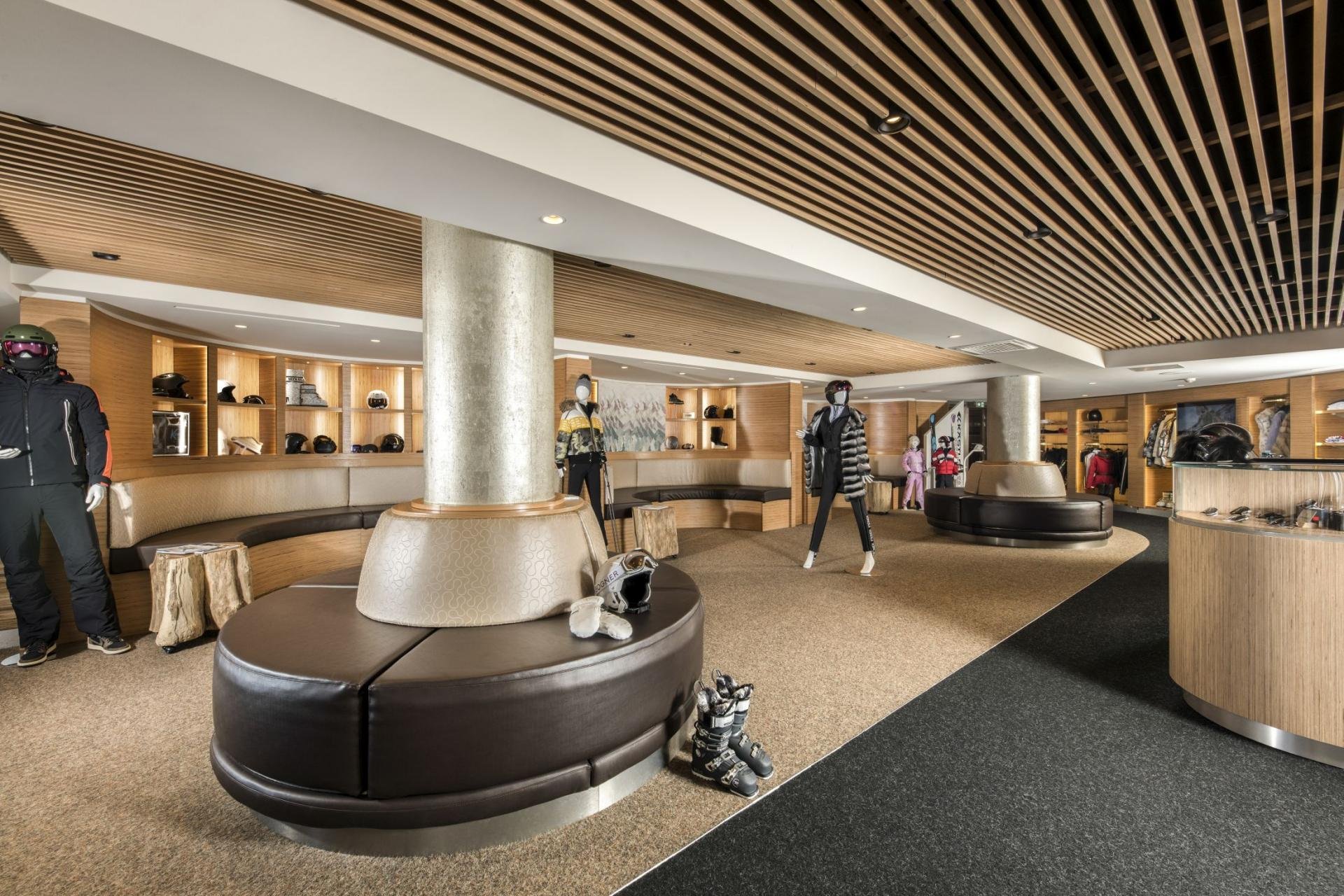 Interior of a retail store with mannequins dressed in winter clothing, shelves with helmets, and seating areas. Modern ceiling design with wooden slats and spotlights.