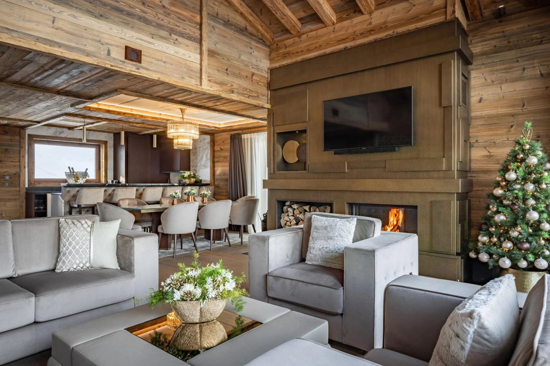 Open-concept living room and dining area in a rustic-style home with wood-paneled walls and ceiling, decorated for Christmas with a Christmas tree. Features include a fireplace with a TV mounted above, a beige sofa, a glass-topped coffee table with a