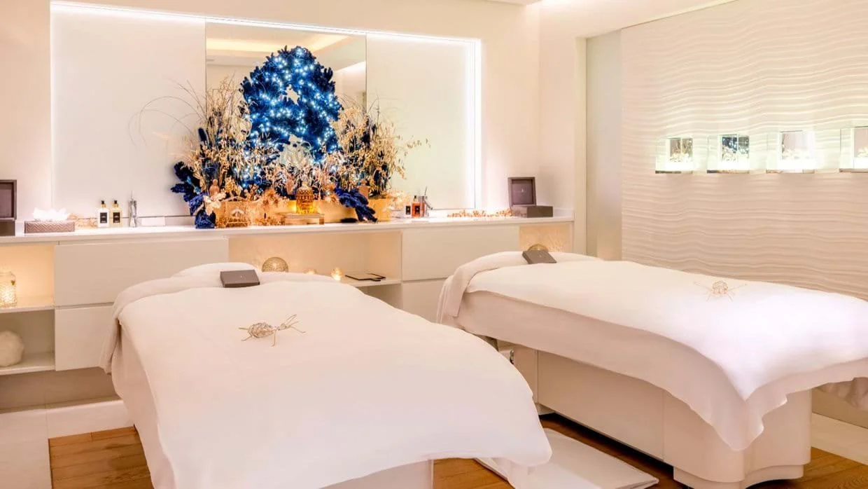 Spa treatment room with two massage beds draped in white sheets, decorated foamy with small decor, with festive Christmas decor including a Christmas tree with lights and ornaments behind, and various candles and treatments on side tables.