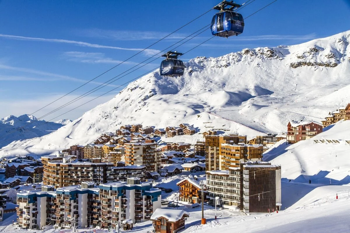 Cable cars traveling above a snow-covered Val Thorens ski resort with multi-story buildings and mountainous terrain in the background.