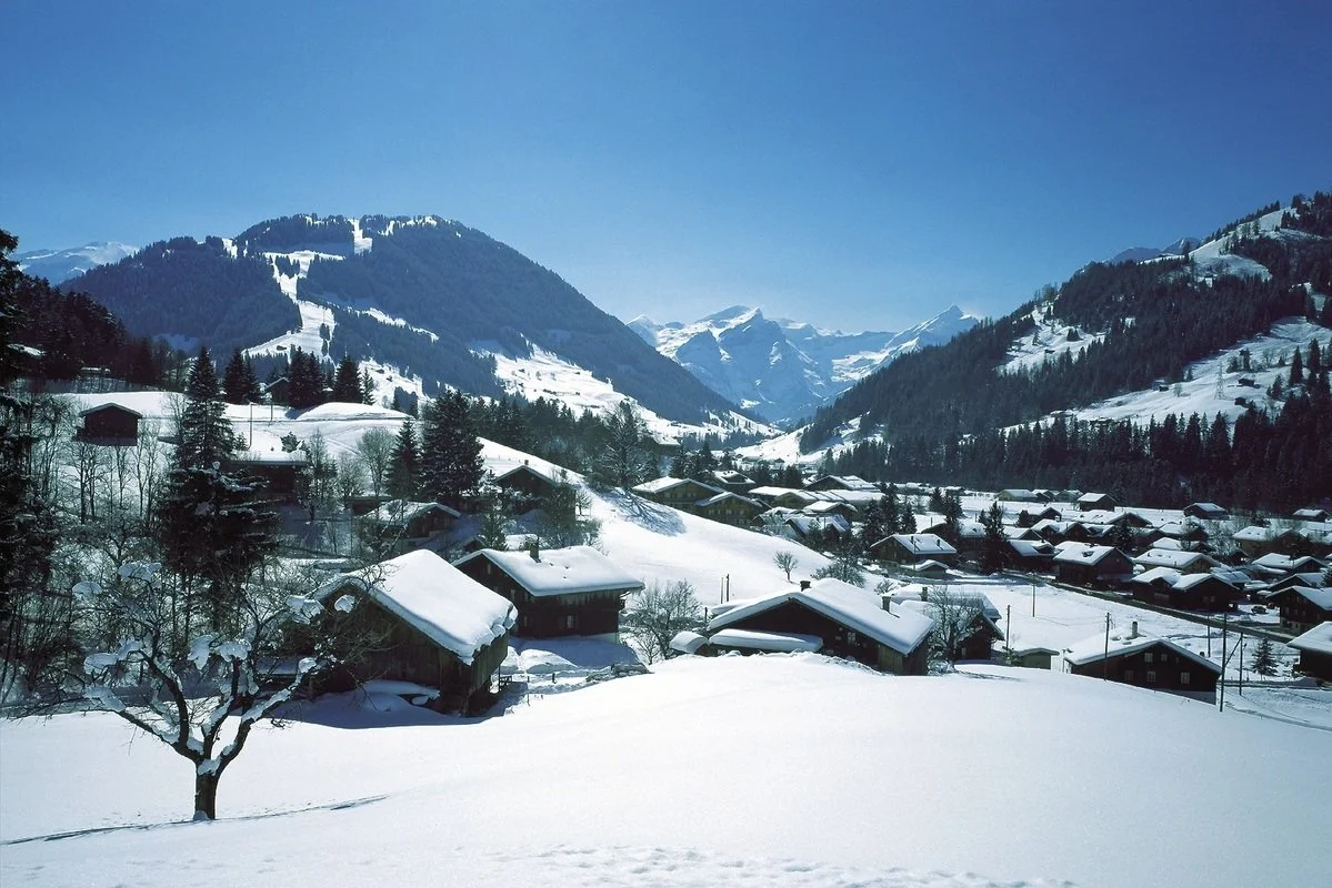Snow-covered Gstaad village in a mountain valley with ski slopes, under a clear blue sky.