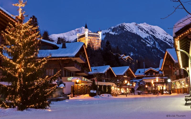 Snow-covered Gstaad village street at dusk with lit Christmas lights and decorated Christmas tree, mountain backdrop with a castle-like building on a hill.