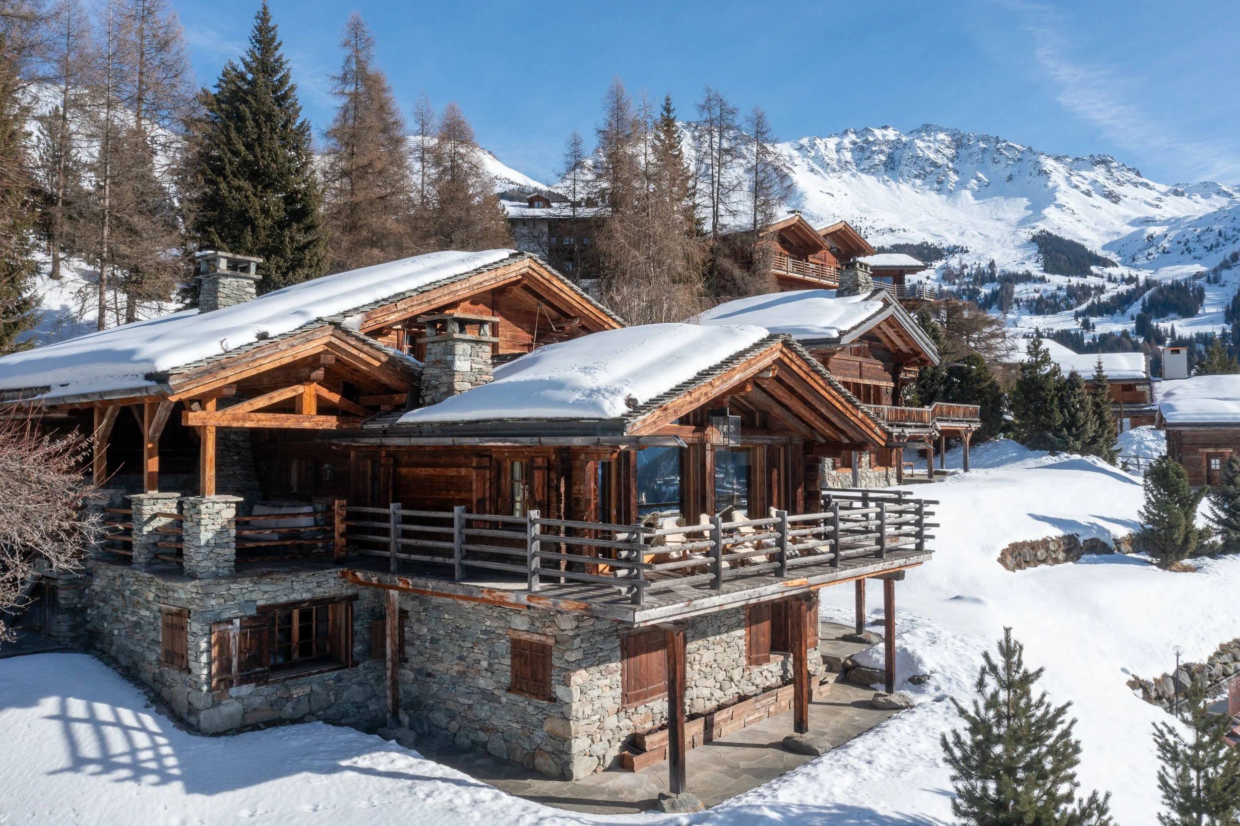 Snow-covered chalet-style wooden houses with stone accents and fenced patios in a mountain landscape, with snow-covered trees and tall snow-capped mountains in the background under a blue sky.