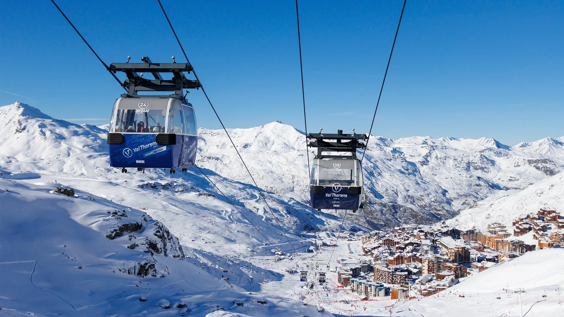 Ski gondolas suspended over a snow-covered Val Thorens mountain landscape with a village below and mountains in the background.