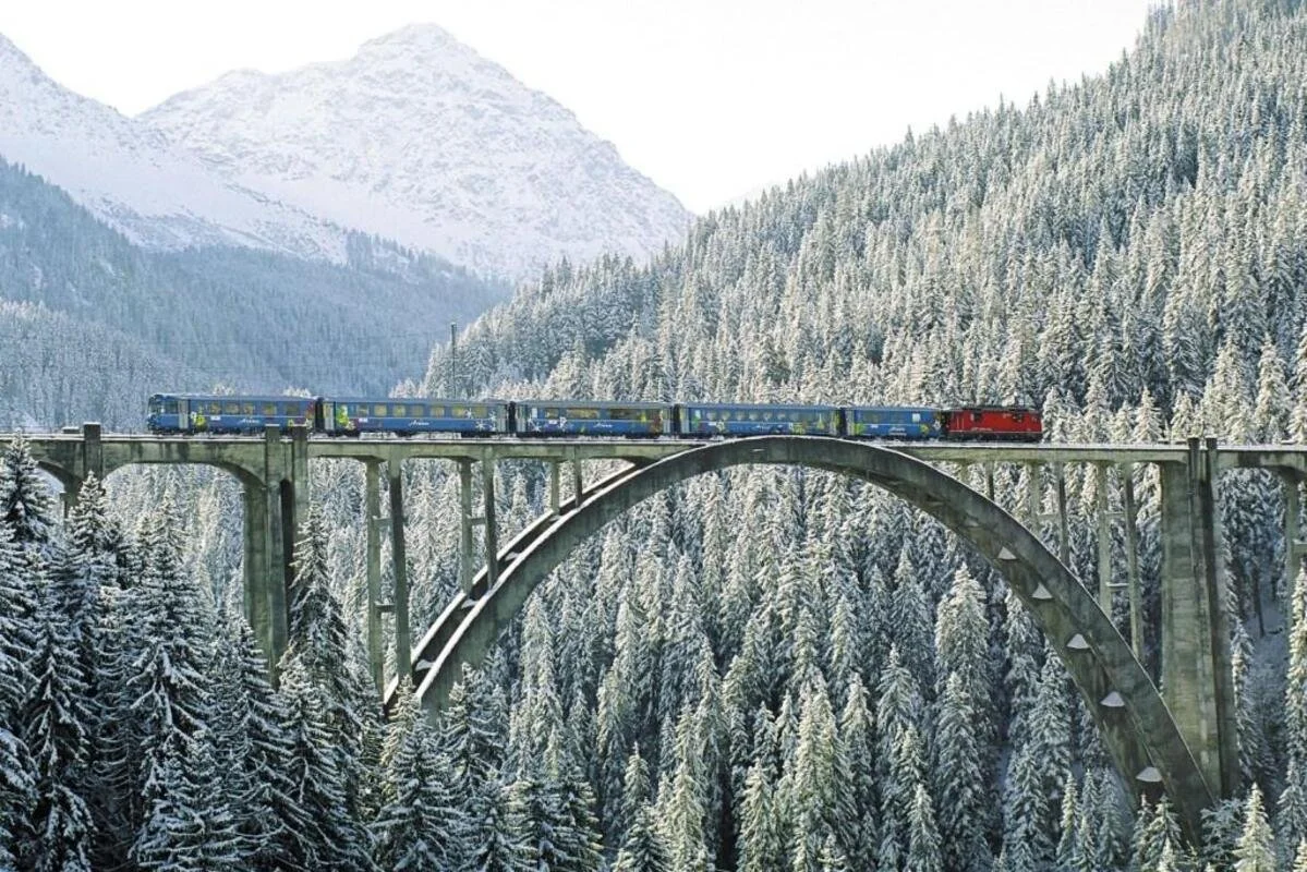 A train with blue and yellow carriages and a red locomotive crossing a tall stone arch bridge over a snow-covered forest in Switzerland with mountains in the background.