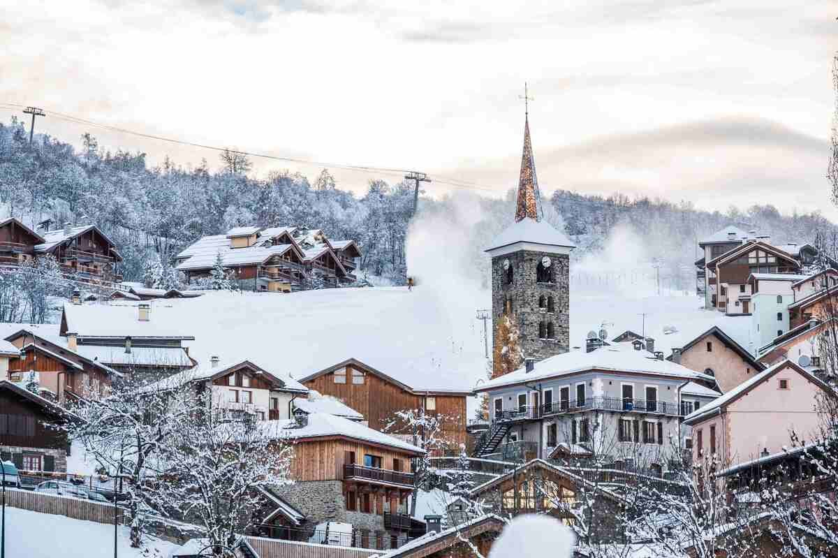 Snow-covered St Martin de Belleville village with a church steeple in the center, surrounded by wooden houses and trees, on a hillside during winter.