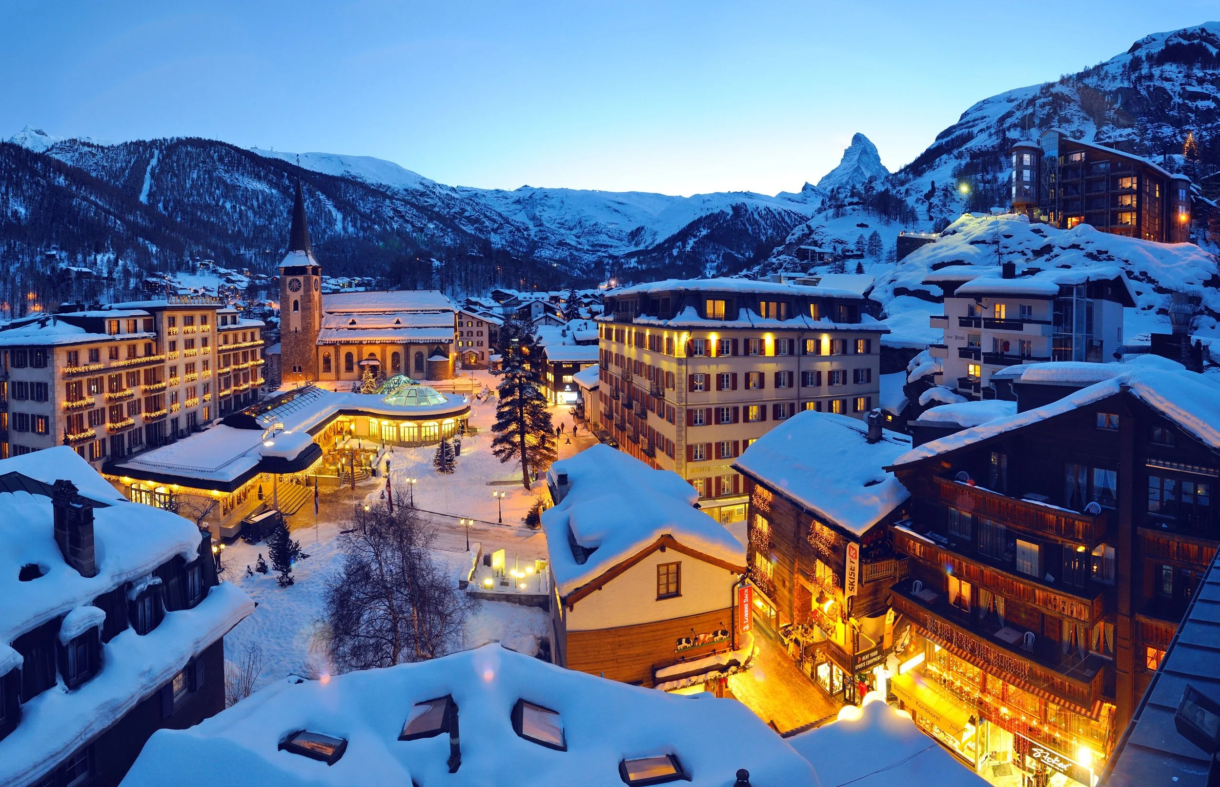 A snowy Zermatt mountain village during dusk with illuminated buildings and a church in the center, surrounded by snow-covered mountains.