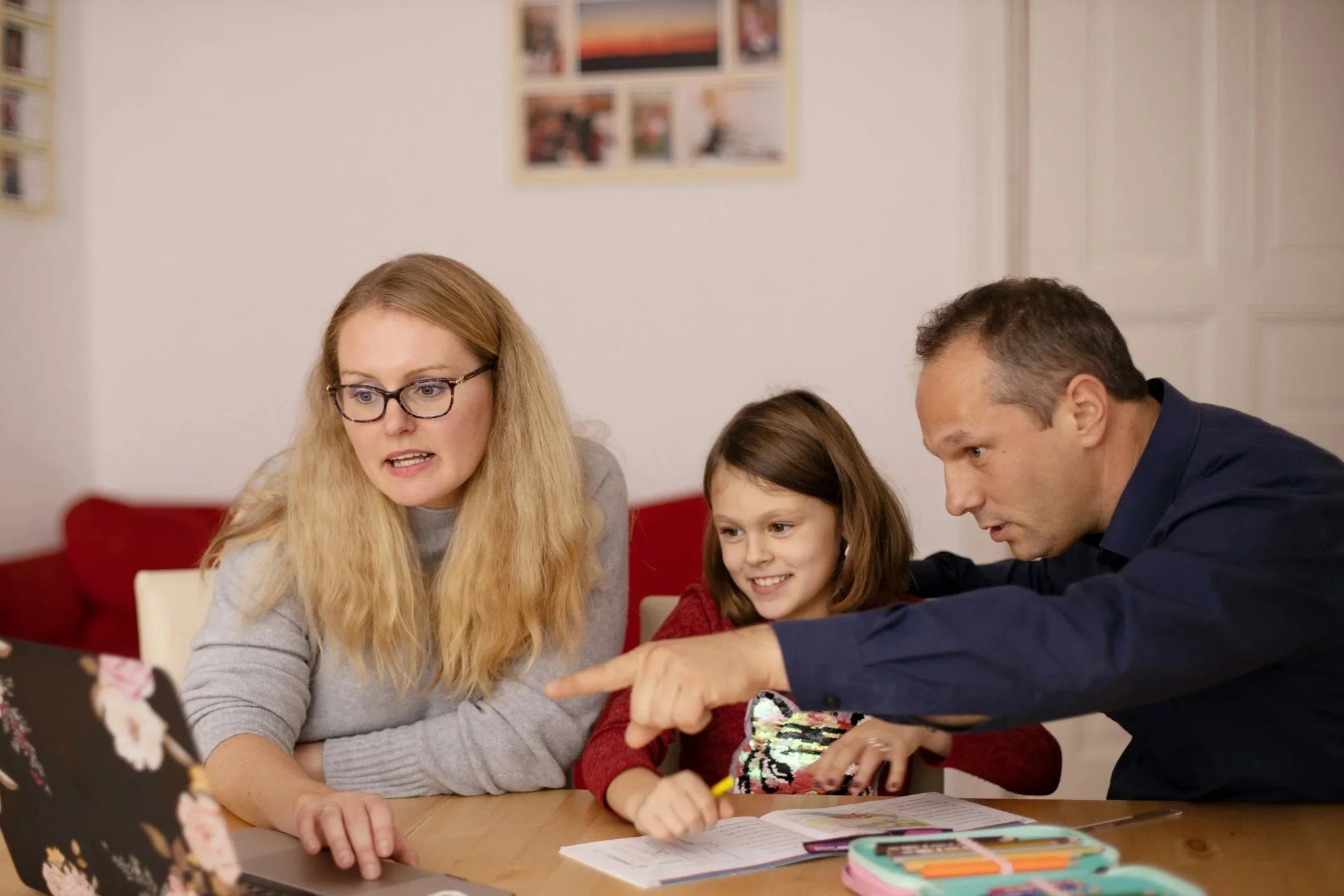 A family of four, including a woman with long blonde hair and glasses, a man, and two young girls, are gathered around a table, looking at a laptop and a colorful gift bag, with papers and stationery on the table.
