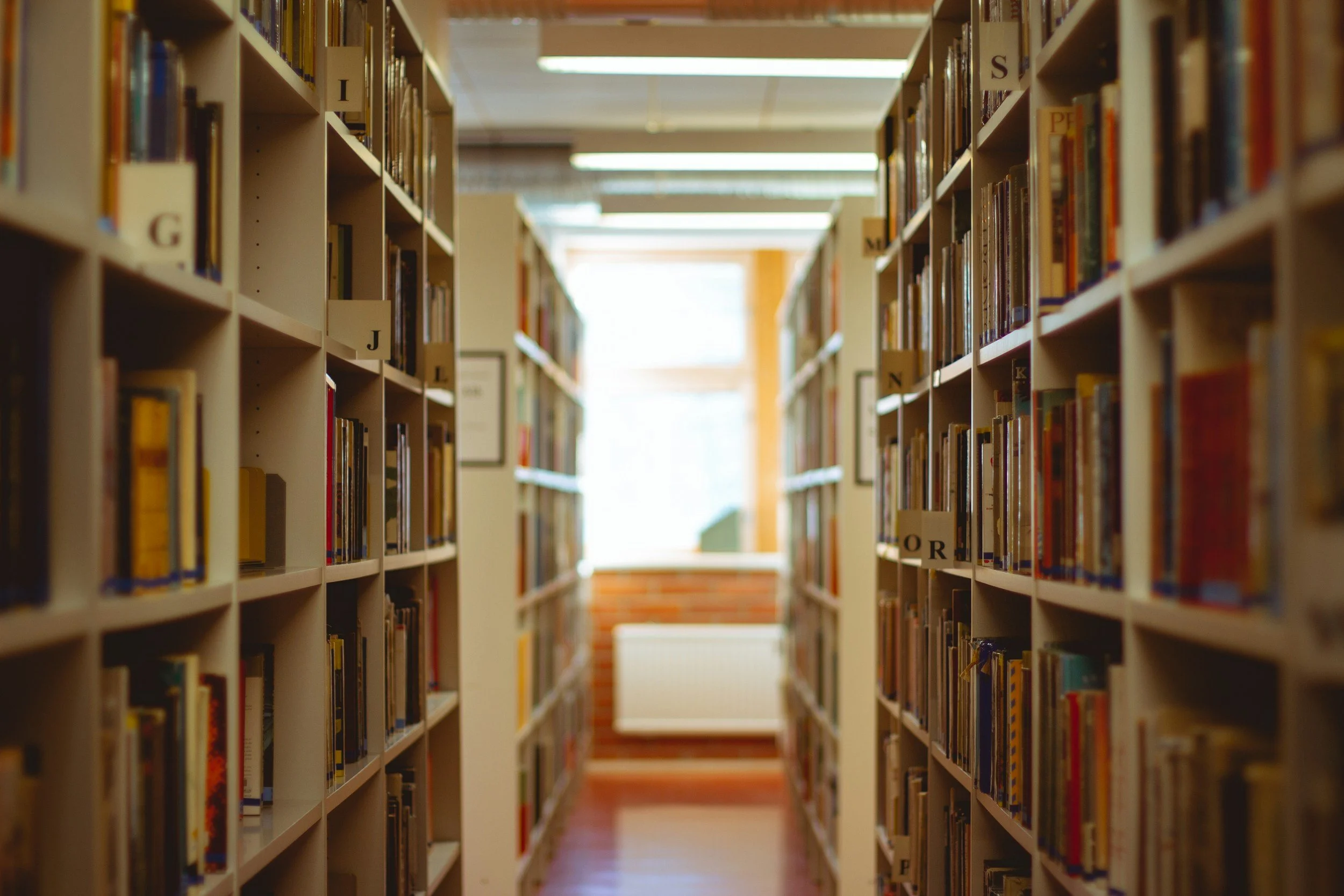 View of bookshelves in a library aisle, filled with books, with a destination at the end of the aisle illuminated by natural light.