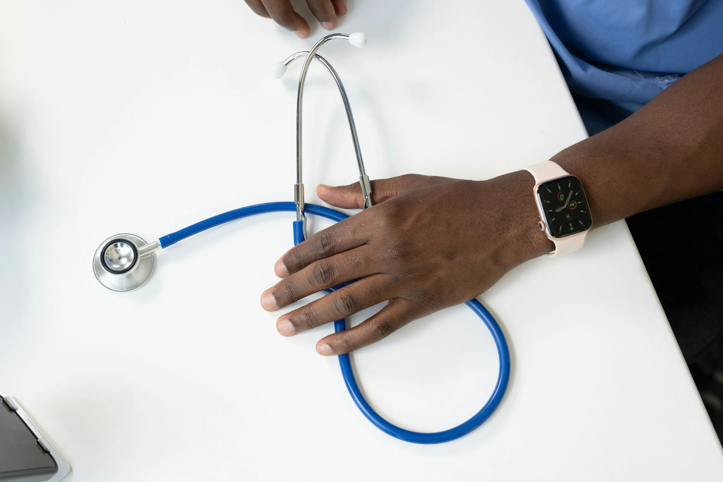 A person with dark skin wearing a smartwatch and a blue scrubs sleeve, resting their hand on a white surface, with a blue stethoscope lying on the surface.