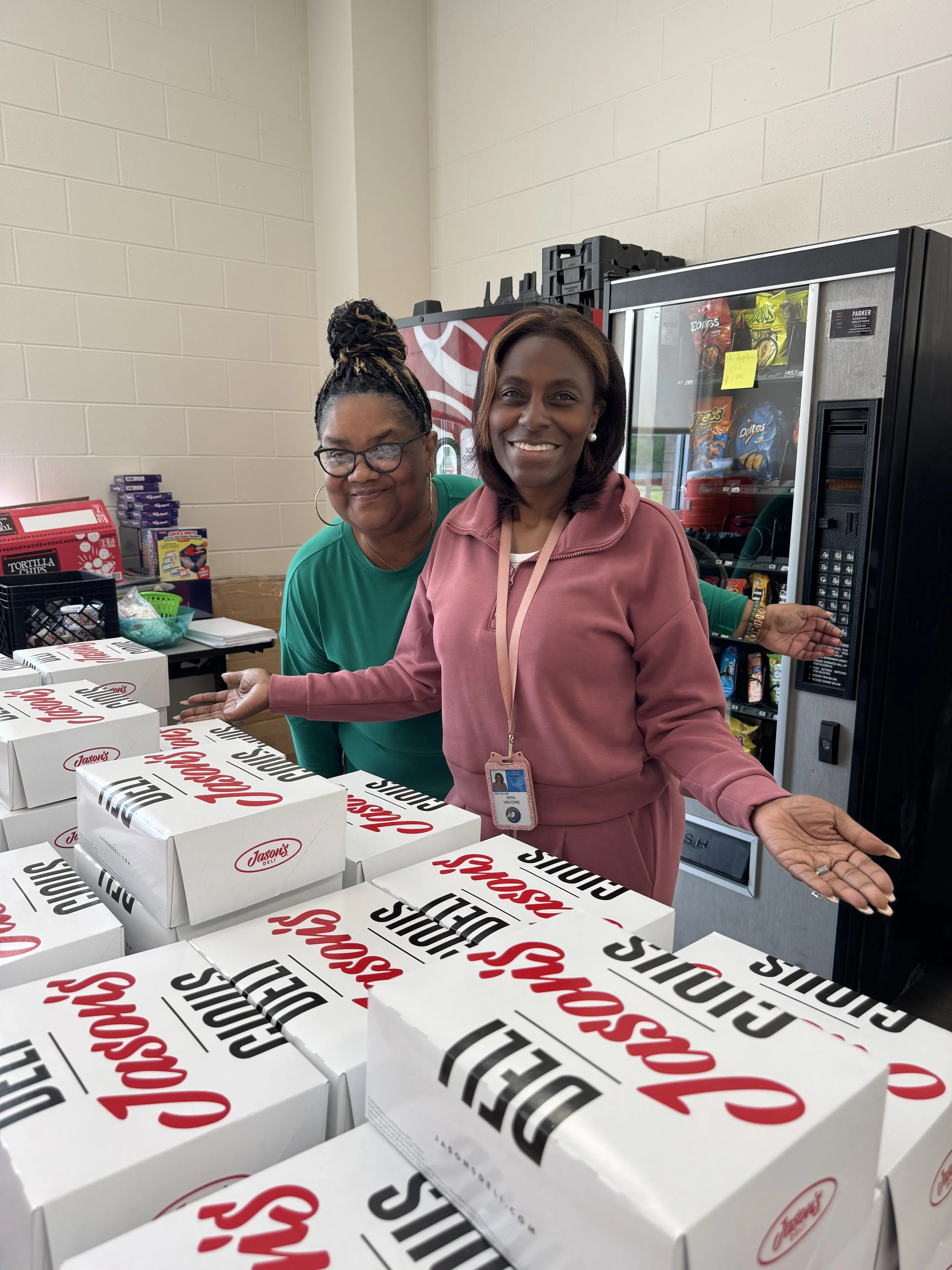 Two women smiling at a table with stacked pizza boxes from Jason's Deli, with a vending machine in the background.