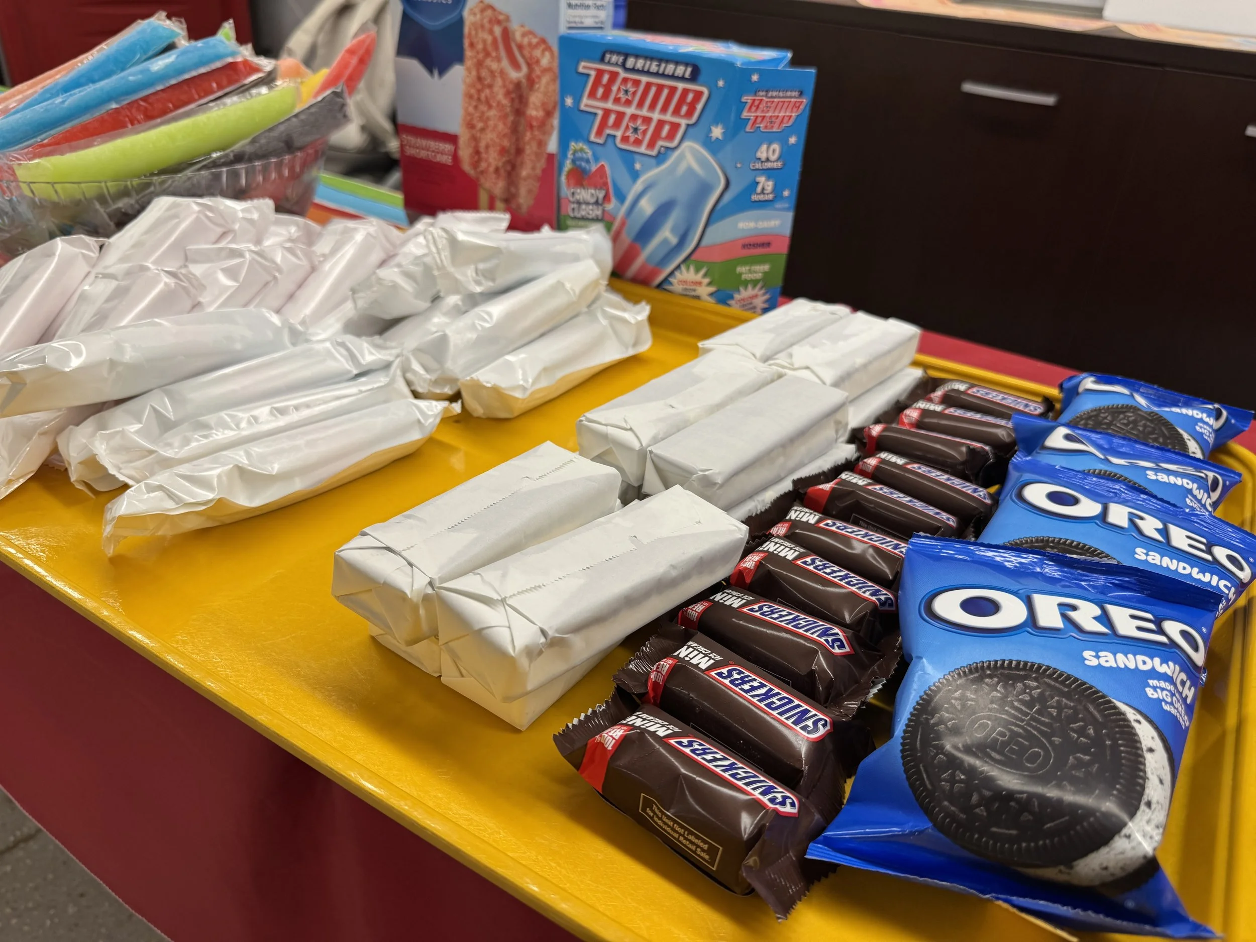 Assorted candy and snack items on a yellow display table. Visible products include candy bars, Oreo cookies, and a box of Bomb Pop ice pops. Some items are partially wrapped or in packaging.