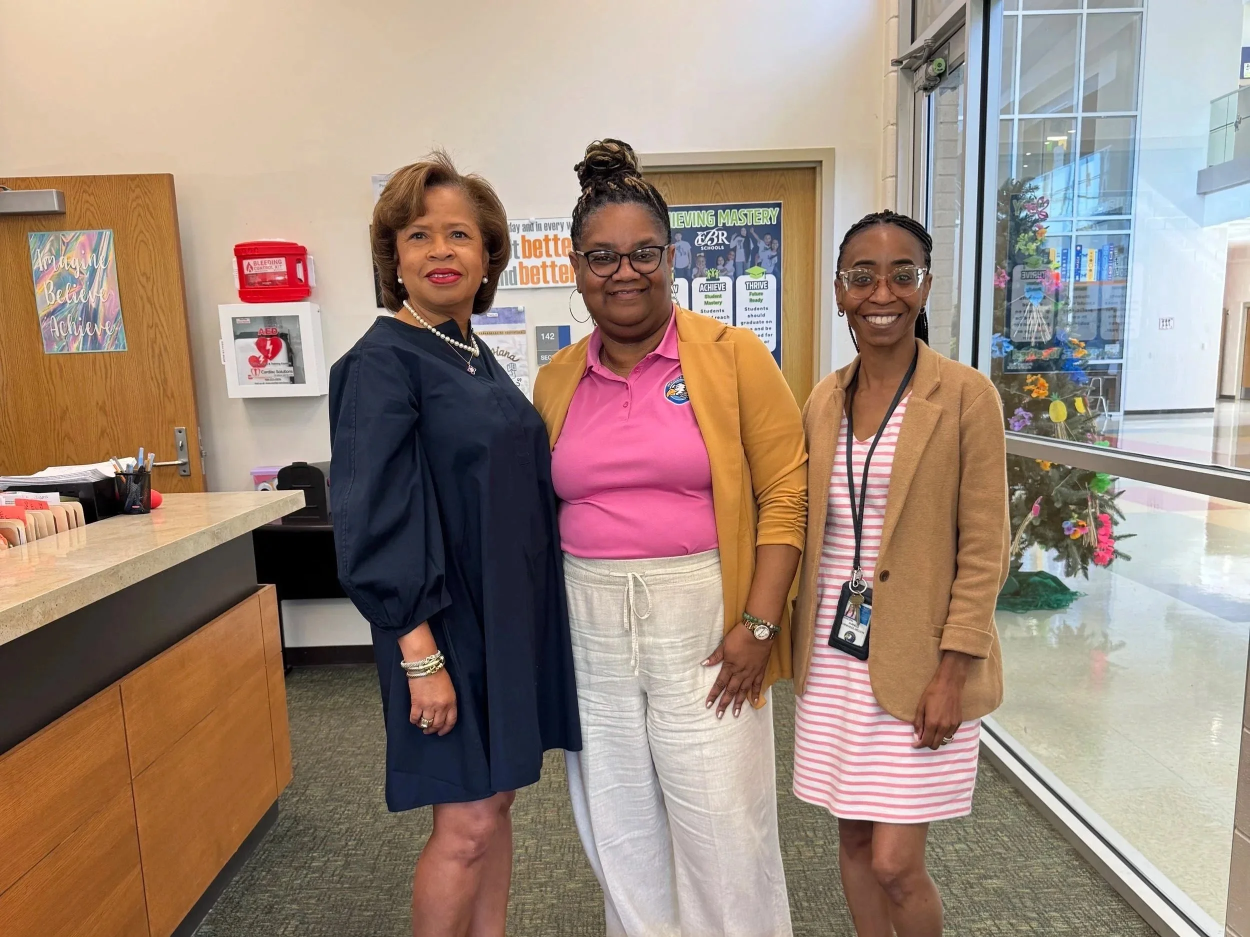 Three women standing together inside a building, smiling at the camera. The woman on the left is wearing a navy dress and pearl jewelry, the woman in the middle is wearing a pink shirt and white pants, and the woman on the right is wearing glasses, a tan blazer, and a striped dress. Behind them, there are posters on the wall, a door with the room number 142, a fire safety sign, and a glass door with a view of plants and a colorful display outside.