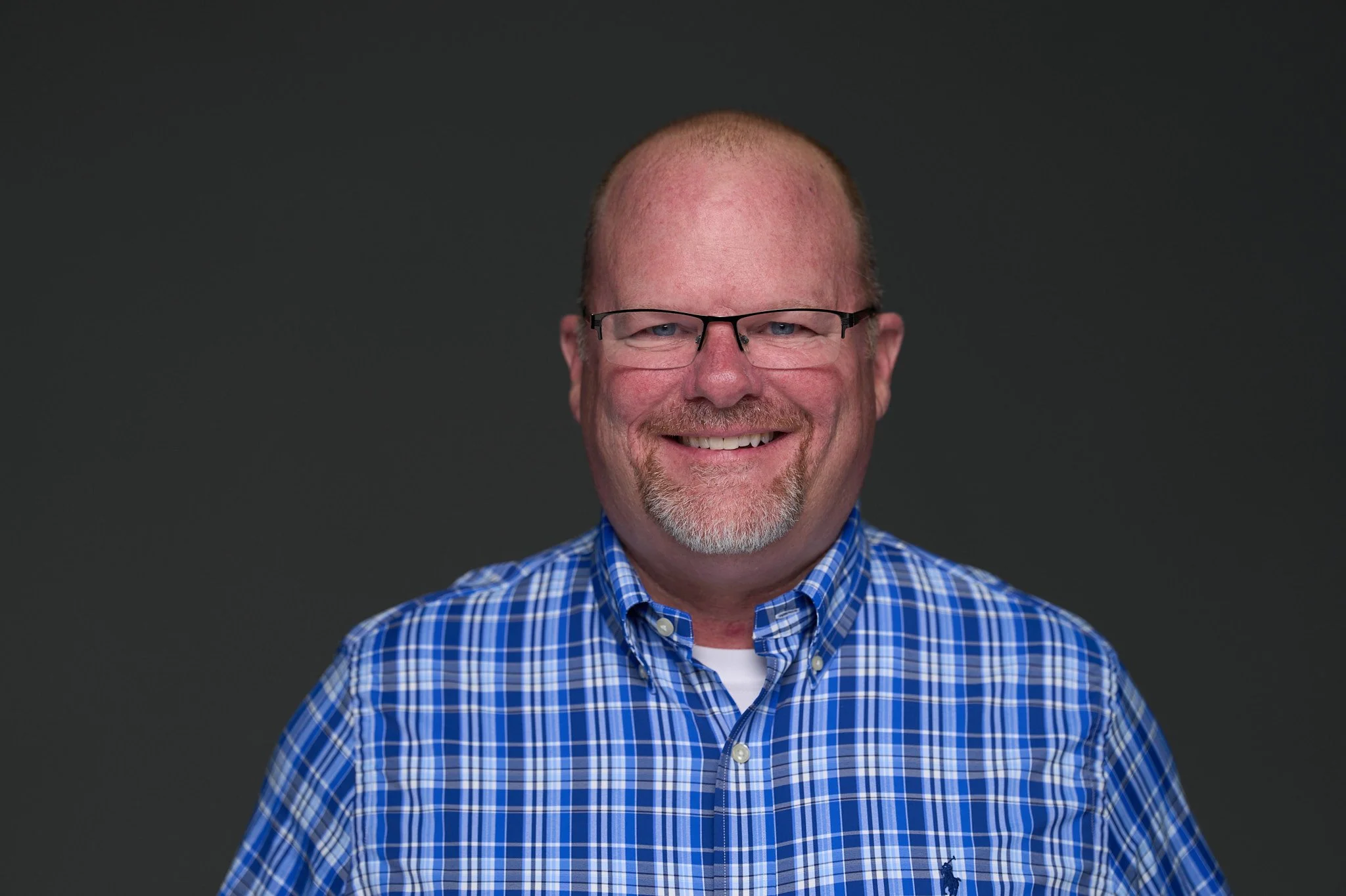 A smiling man with a shaved head, beard, and blue eyes, wearing a colorful plaid shirt, standing outdoors with a blurred background of trees and sky.
