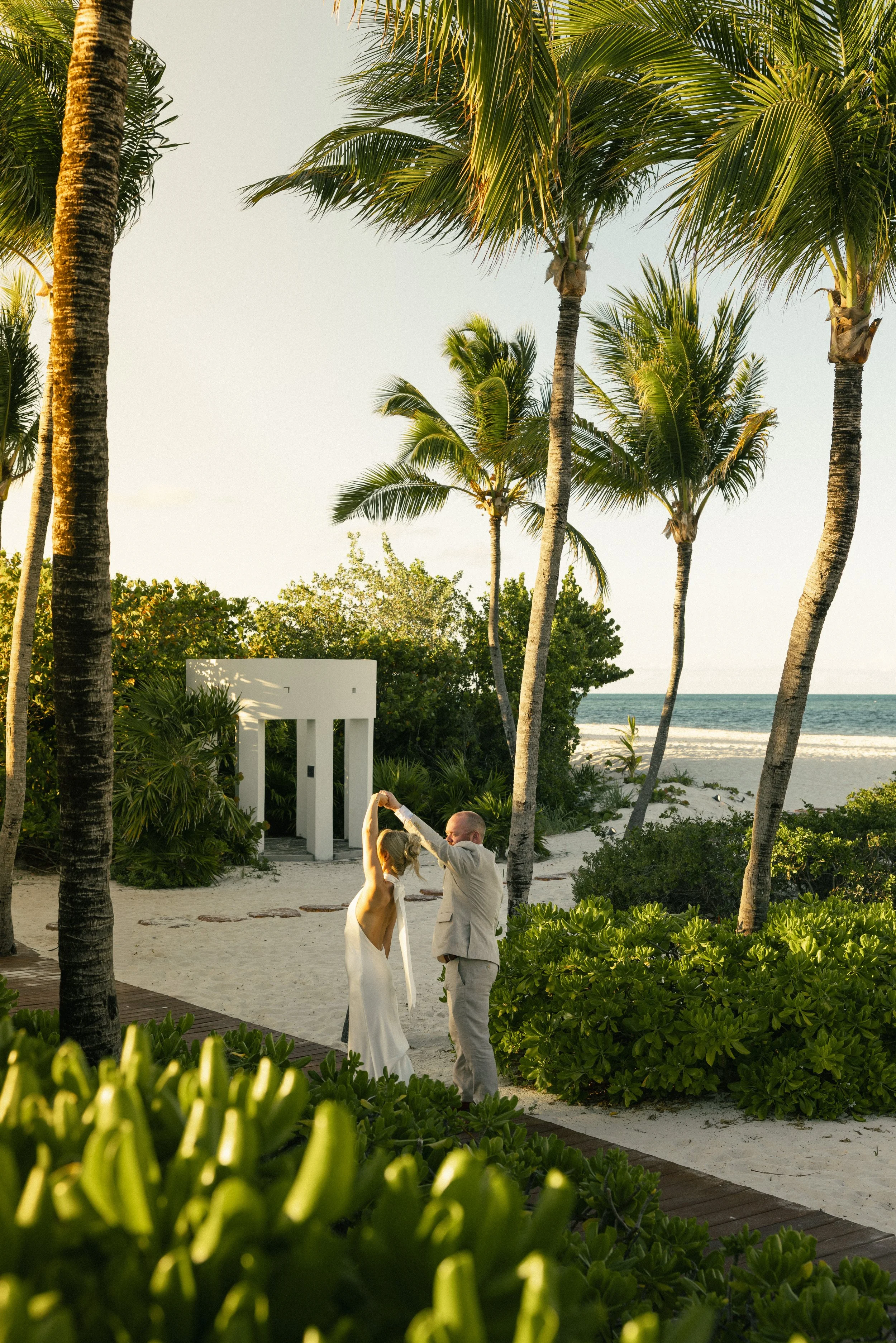 A bride and groom dancing on a sandy beach surrounded by palm trees with the ocean in the background during sunset.