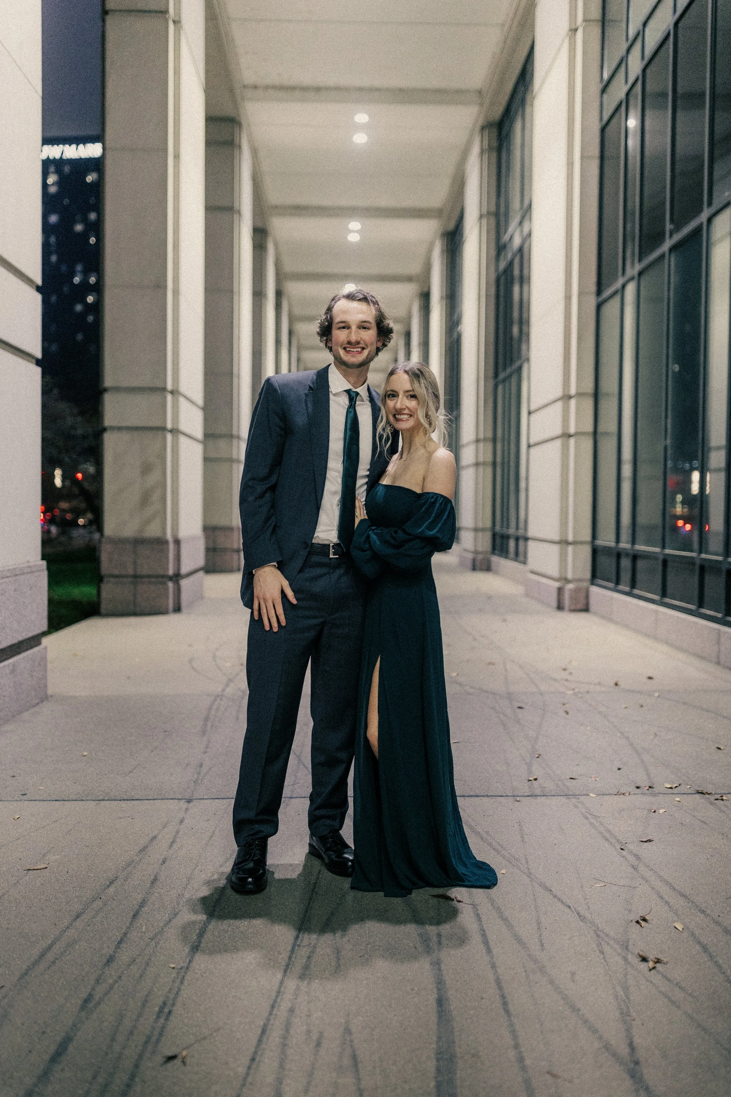 A young man and woman in formal attire standing together under an illuminated covered walkway at night. The man is wearing a suit and tie, and the woman is dressed in an elegant off-the-shoulder gown. They are smiling and posing for the photo.