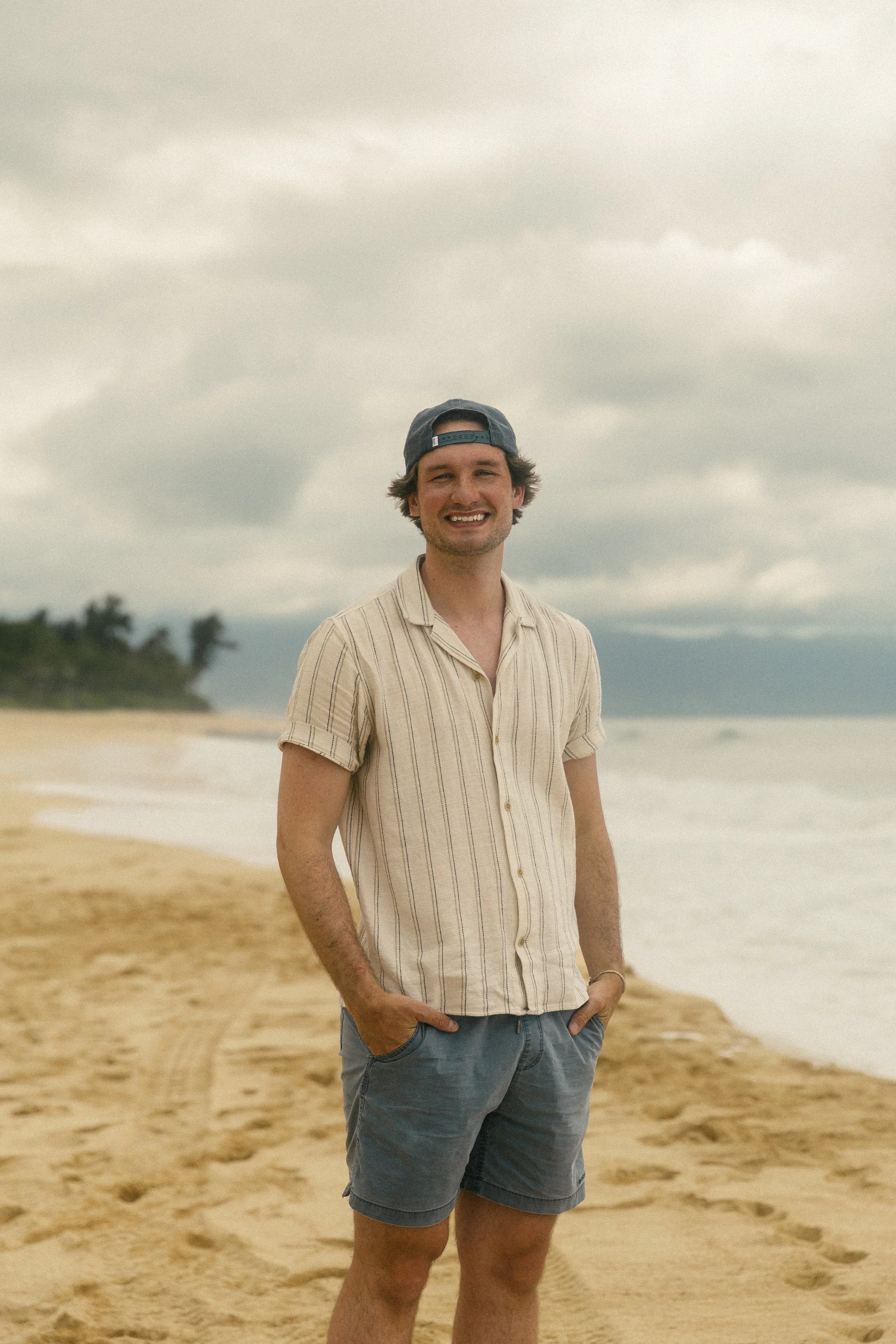 A young man stands on a sandy beach with a cloudy sky overhead, smiling and looking at the camera, wearing a backwards cap, a striped short-sleeved shirt, and shorts.