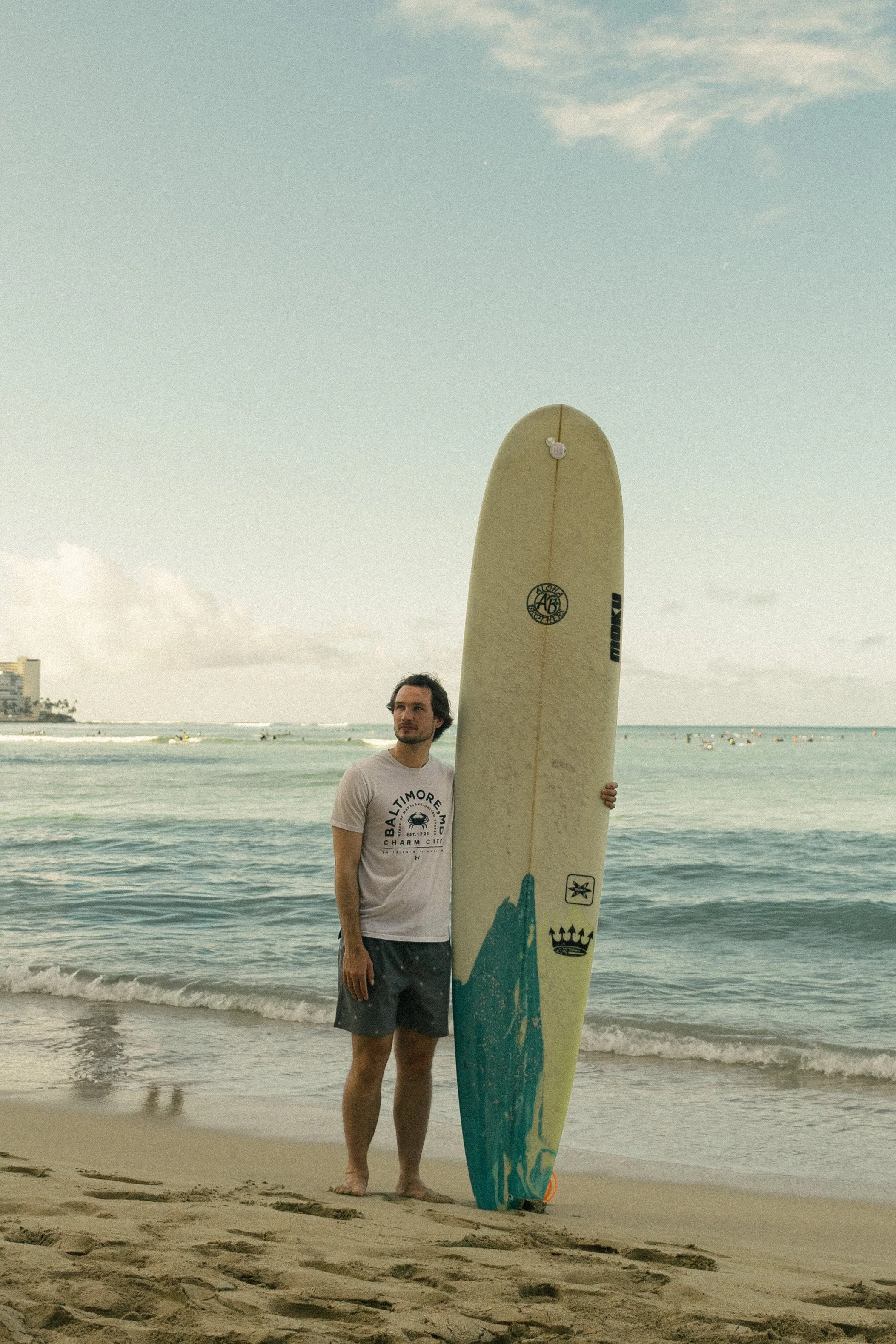 A young man stands on the beach holding a surfboard upright in the sand. The ocean and a few people swimming are visible in the background, with buildings along the coastline and a partly cloudy sky overhead.