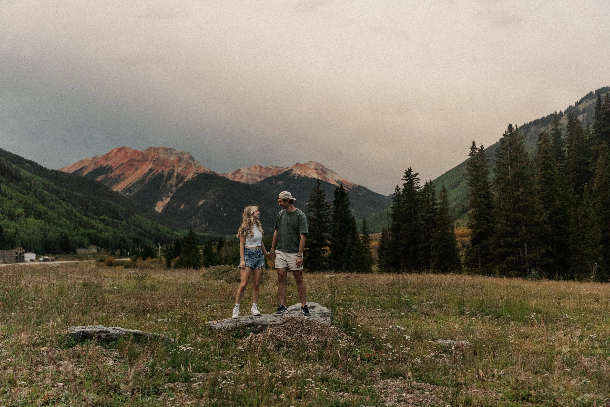 A young couple standing on a large rock in a grassy field with mountains and pine trees in the background, during sunset.