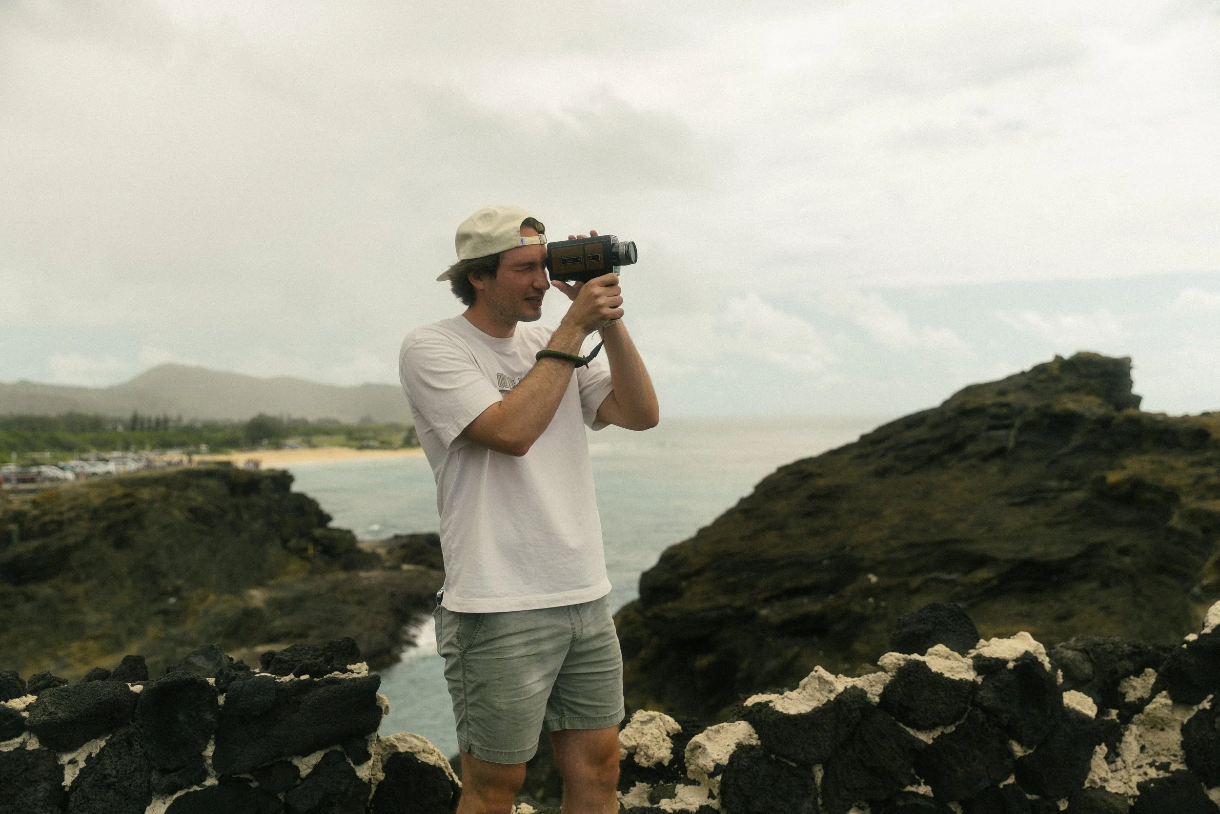 A man wearing a beige cap backwards and a white t-shirt, standing outdoors on rocky terrain, holding a vintage camera to his eye, with an ocean and cloudy sky in the background.