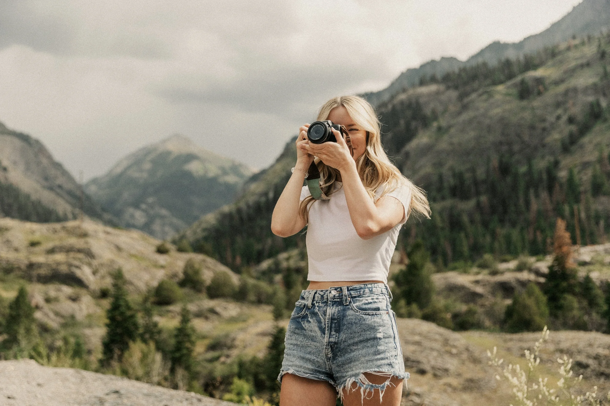 A young blonde woman wearing a white crop top and distressed denim shorts, holding a camera up to her face, standing outdoors with mountainous terrain in the background.