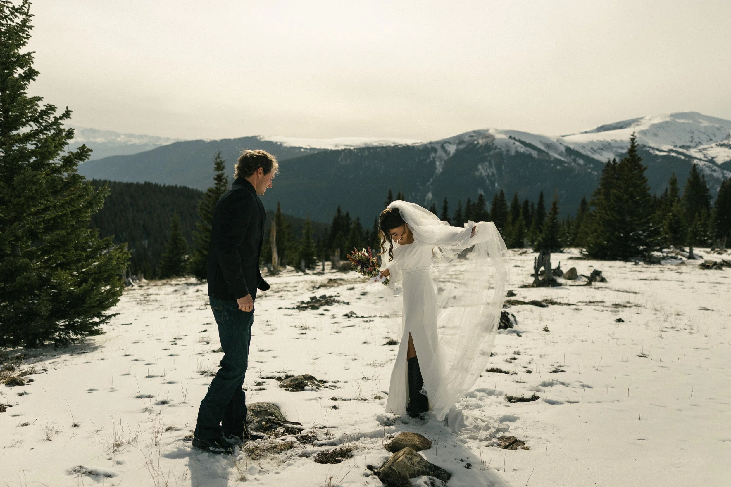 A bride in a white wedding dress with a veil and bouquet is leaning forward slightly, holding her dress, while a groom in a black suit and jeans is standing nearby in a snowy mountain landscape with evergreen trees and snow-capped mountains in the ba