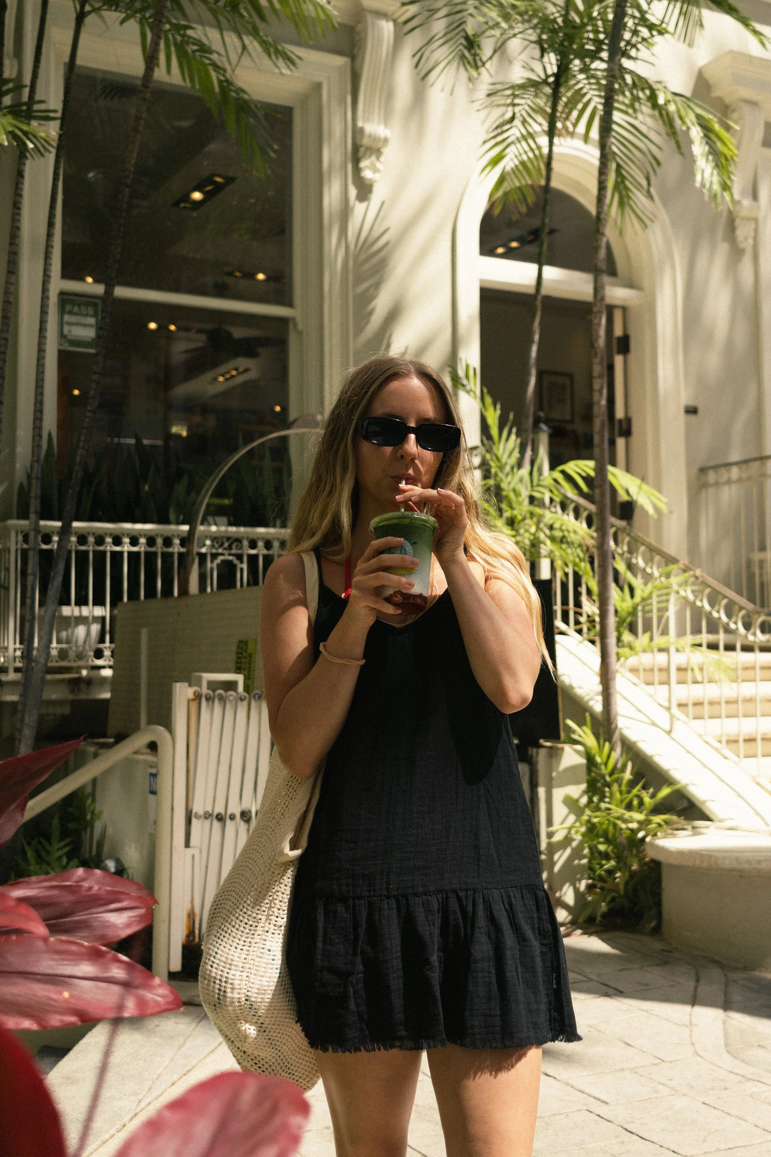 A woman with long hair wearing sunglasses and a black dress is standing outdoors, sipping from a cup with a straw. She also has a beige woven bag hanging from her shoulder. In the background, there are palm trees, a white building with arched windows