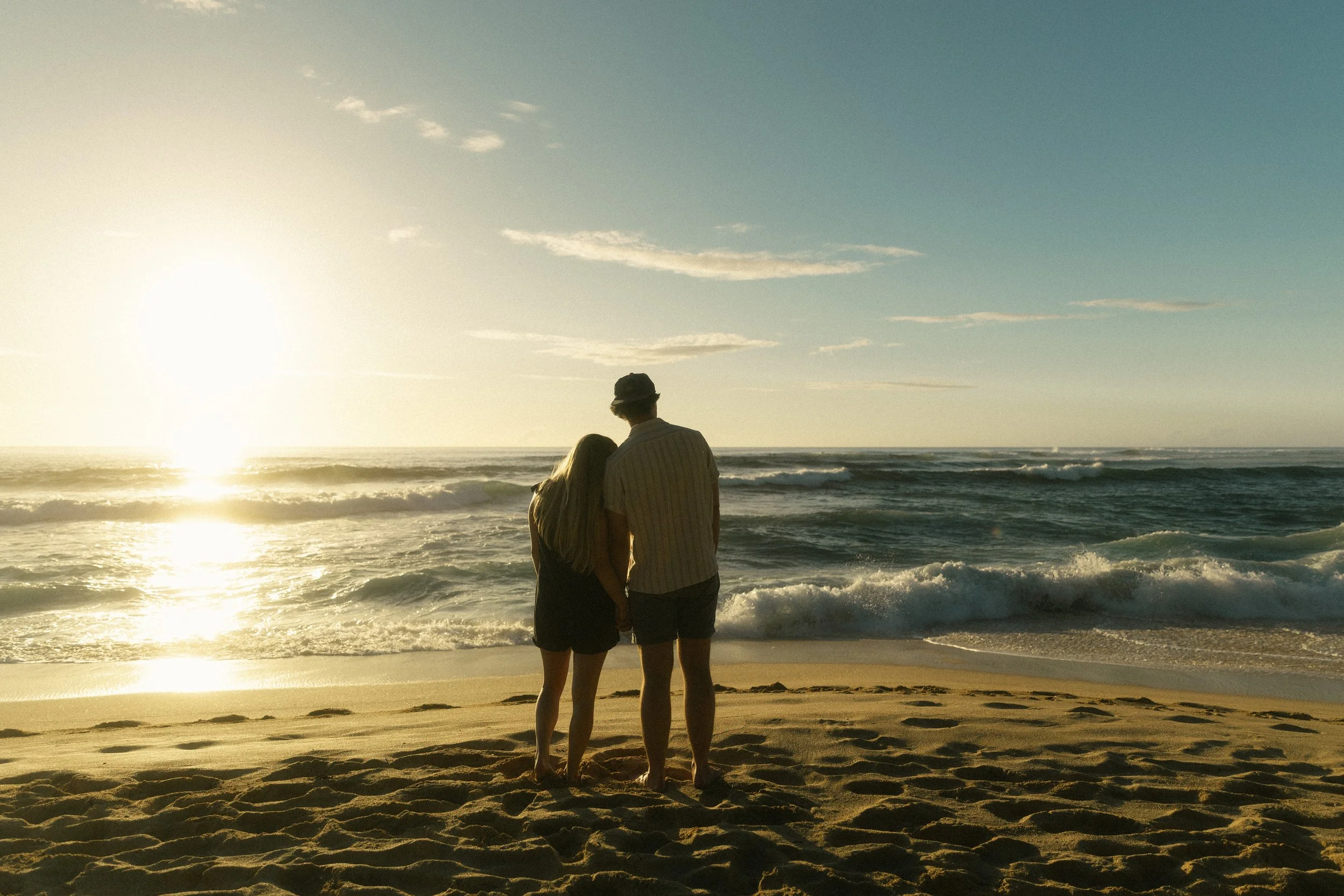 A couple standing on the beach at sunset, overlooking the ocean, with waves crashing and sky partially cloudy.