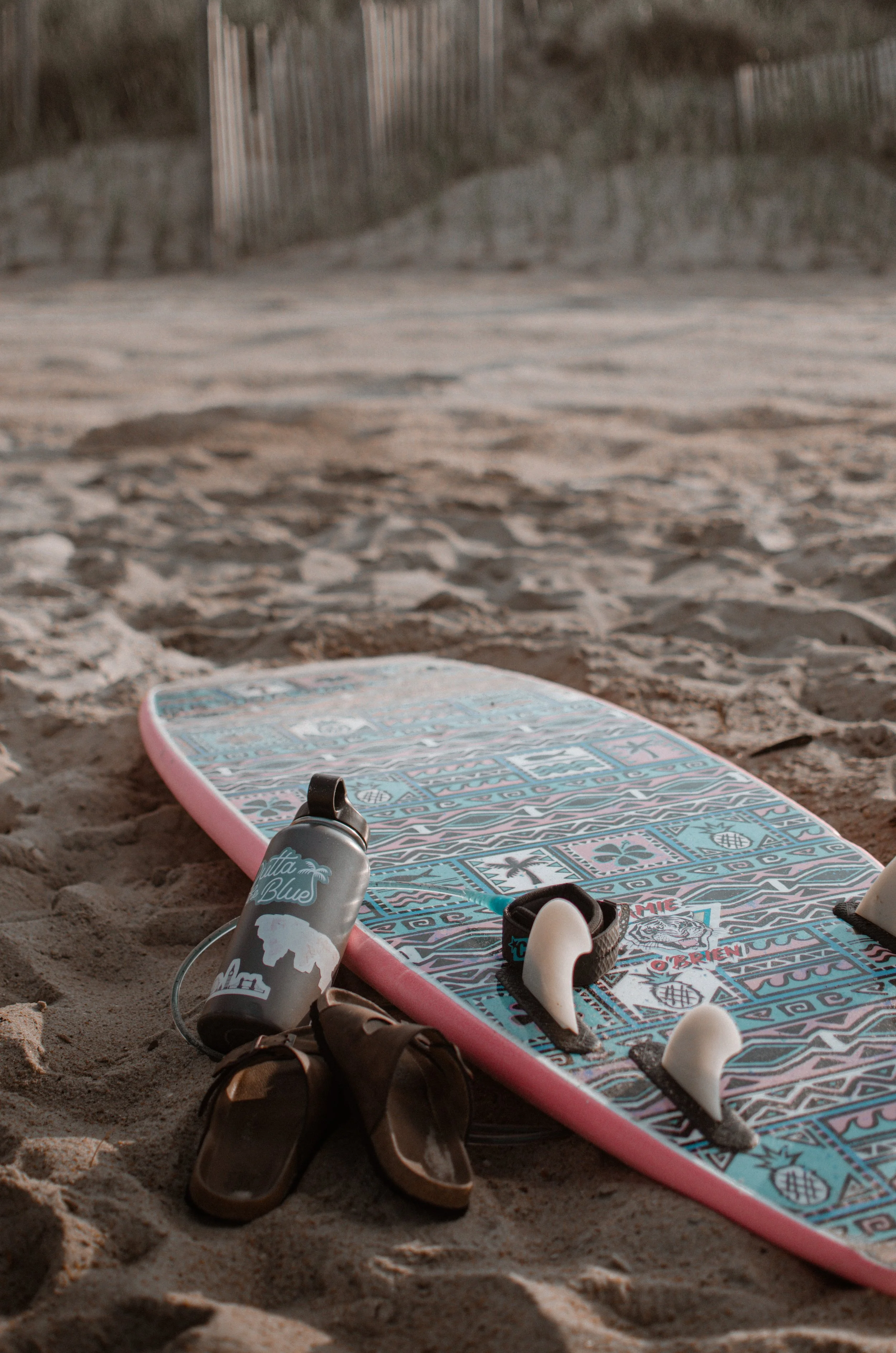 Surfboard with patterned design on sandy beach, with sandals, water bottle, and pair of shoes nearby.