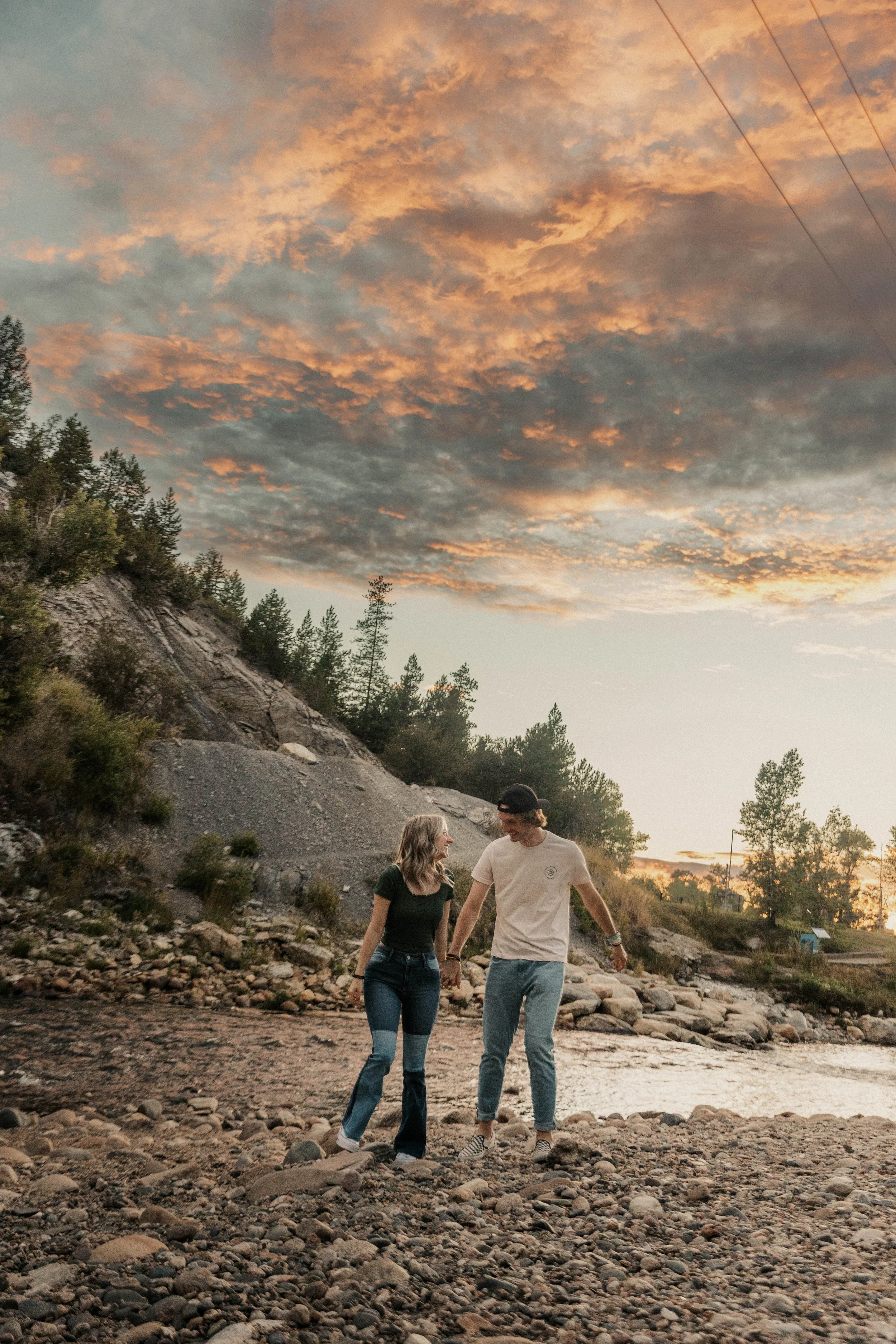 Two young people, a man and a woman, walking hand in hand along a rocky riverbank at sunset. The sky is filled with orange and pink clouds, and there are trees and hills in the background.