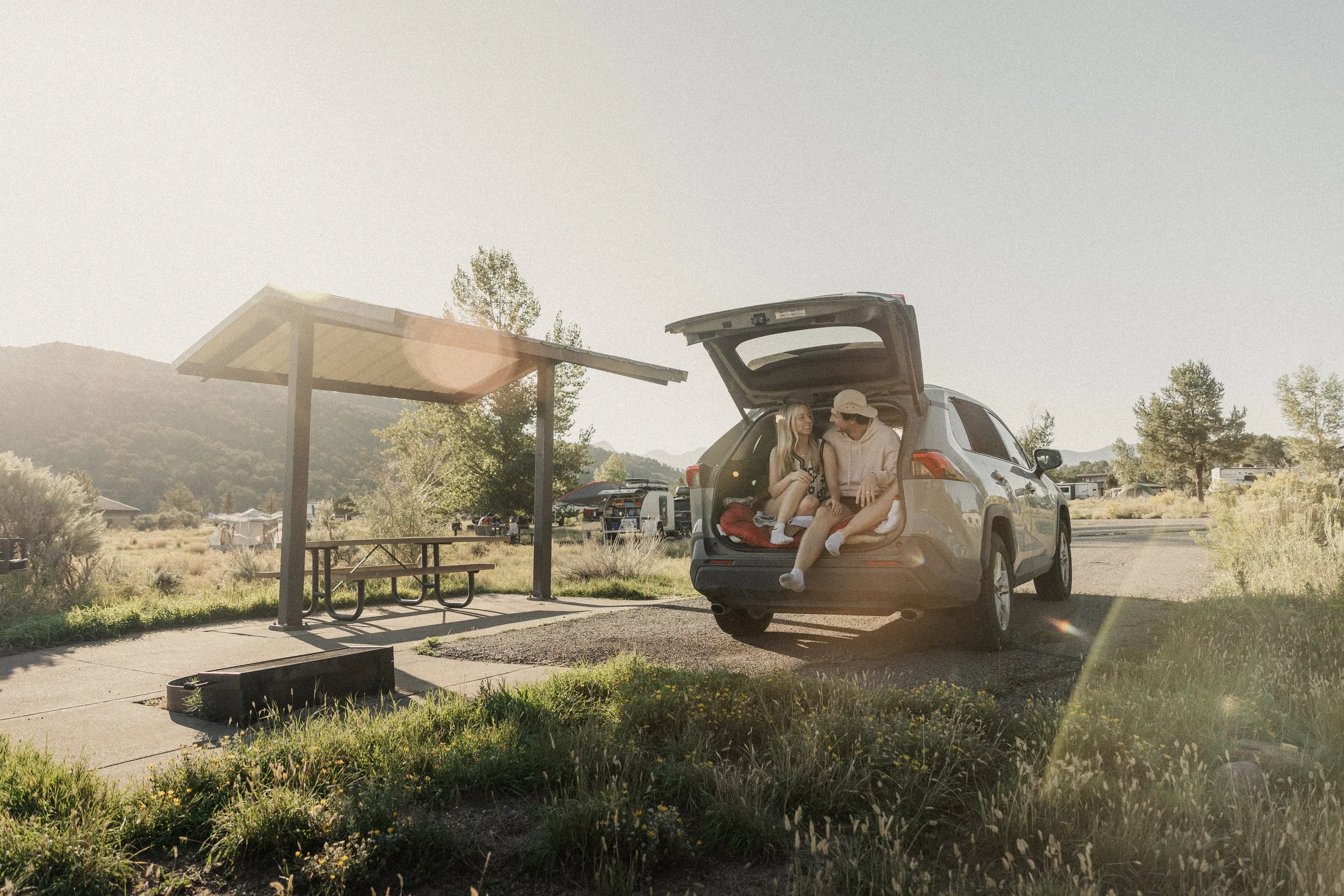 Two people sitting in the open trunk of a parked SUV at a rest area with a picnic table and a solar panel shelter, surrounded by trees and mountains during sunset.