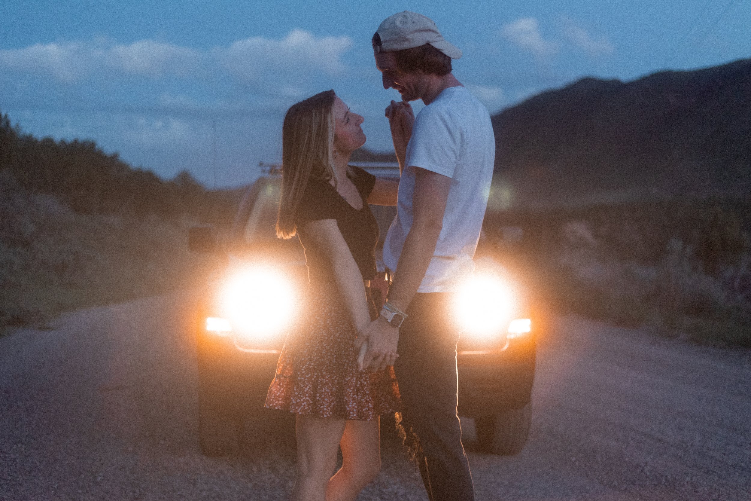 A couple stands close together on a road at dusk, holding hands and smiling at each other, with car headlights behind them and a mountain in the background.