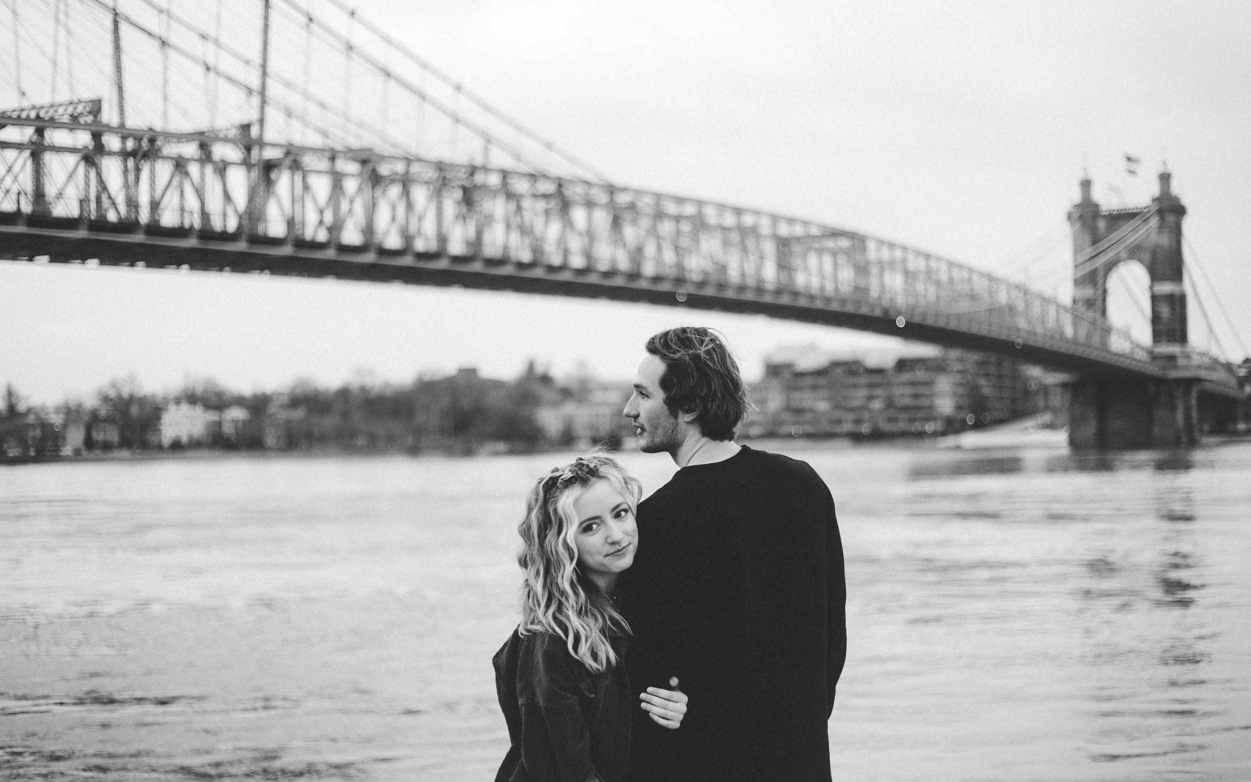A black and white photograph of a man and woman standing near the water with a bridge in the background, possibly the Brooklyn Bridge.