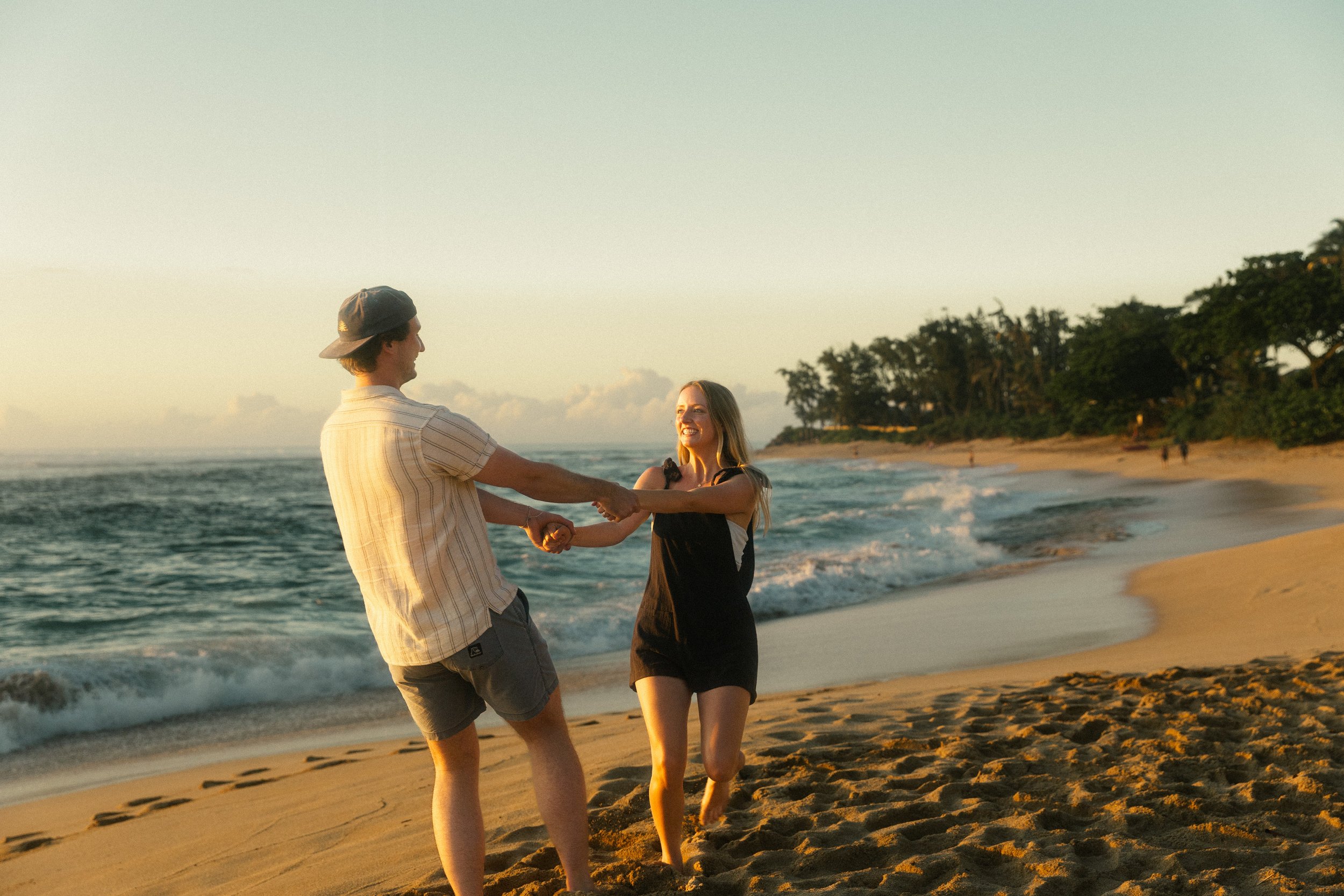 A couple holding hands and dancing on a sandy beach at sunset, with waves crashing and tropical trees in the background.