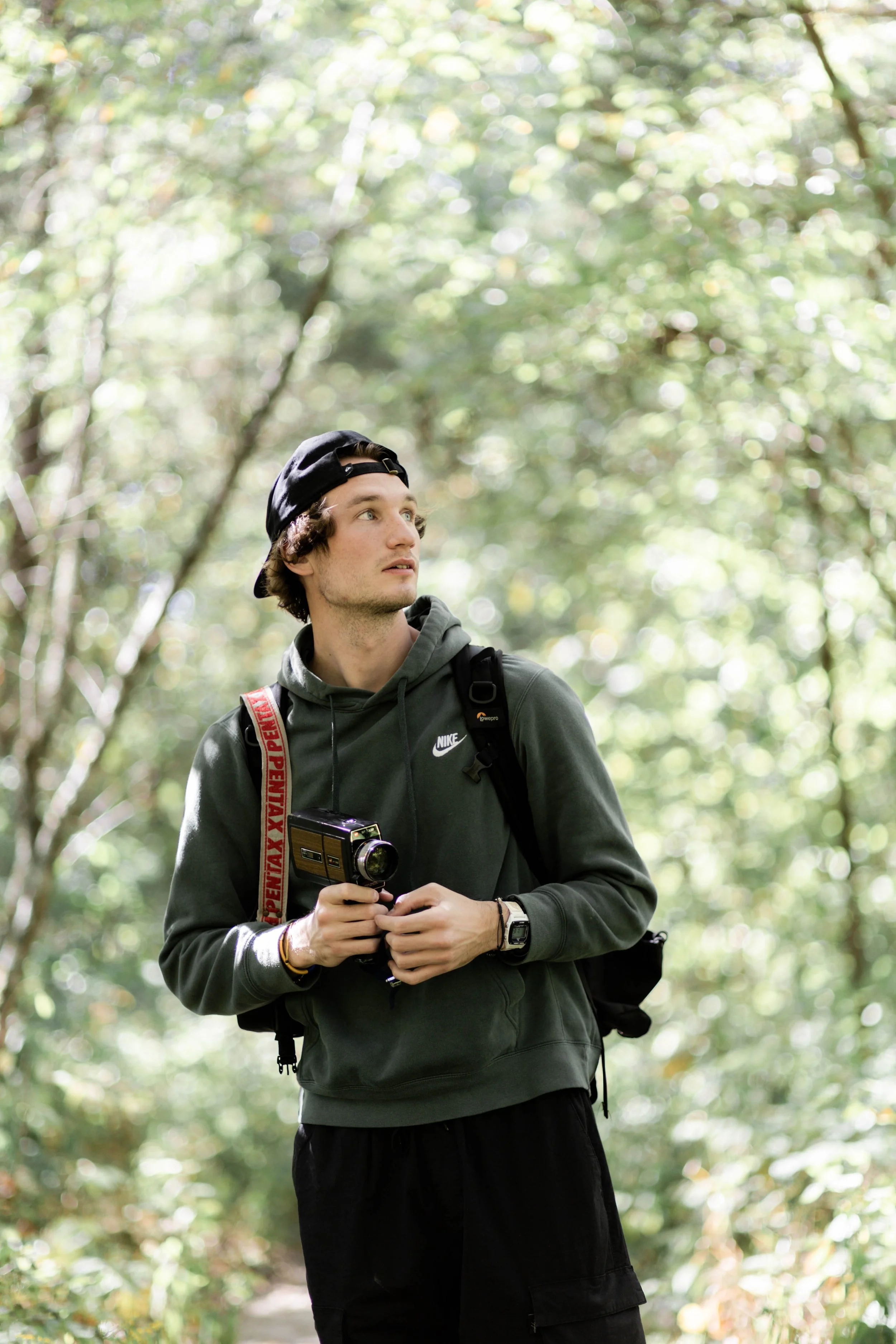 Young man with curly hair wearing a black hoodie, black cap backwards, and carrying a backpack, holding a vintage camera, walking through a green forest.