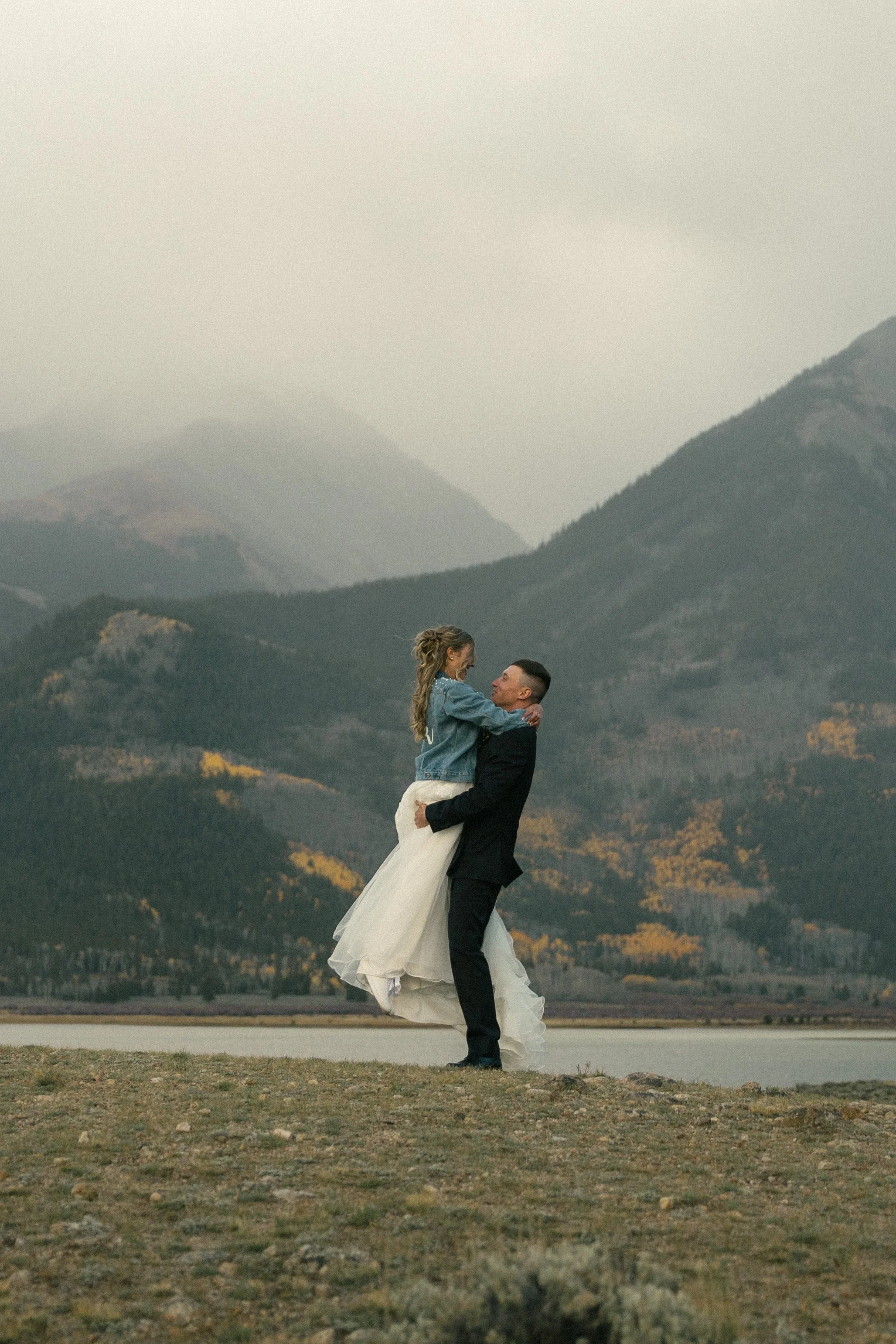A couple dressed in formal wedding attire standing outdoors in a mountainous landscape, with mountains and trees in the background, during daytime.
