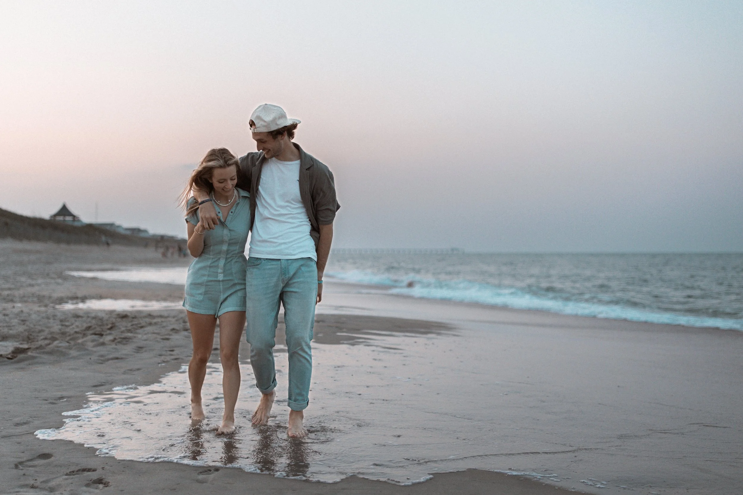 A young couple walking barefoot on the beach at sunset, smiling, with the ocean waves nearby and a pier in the distance.