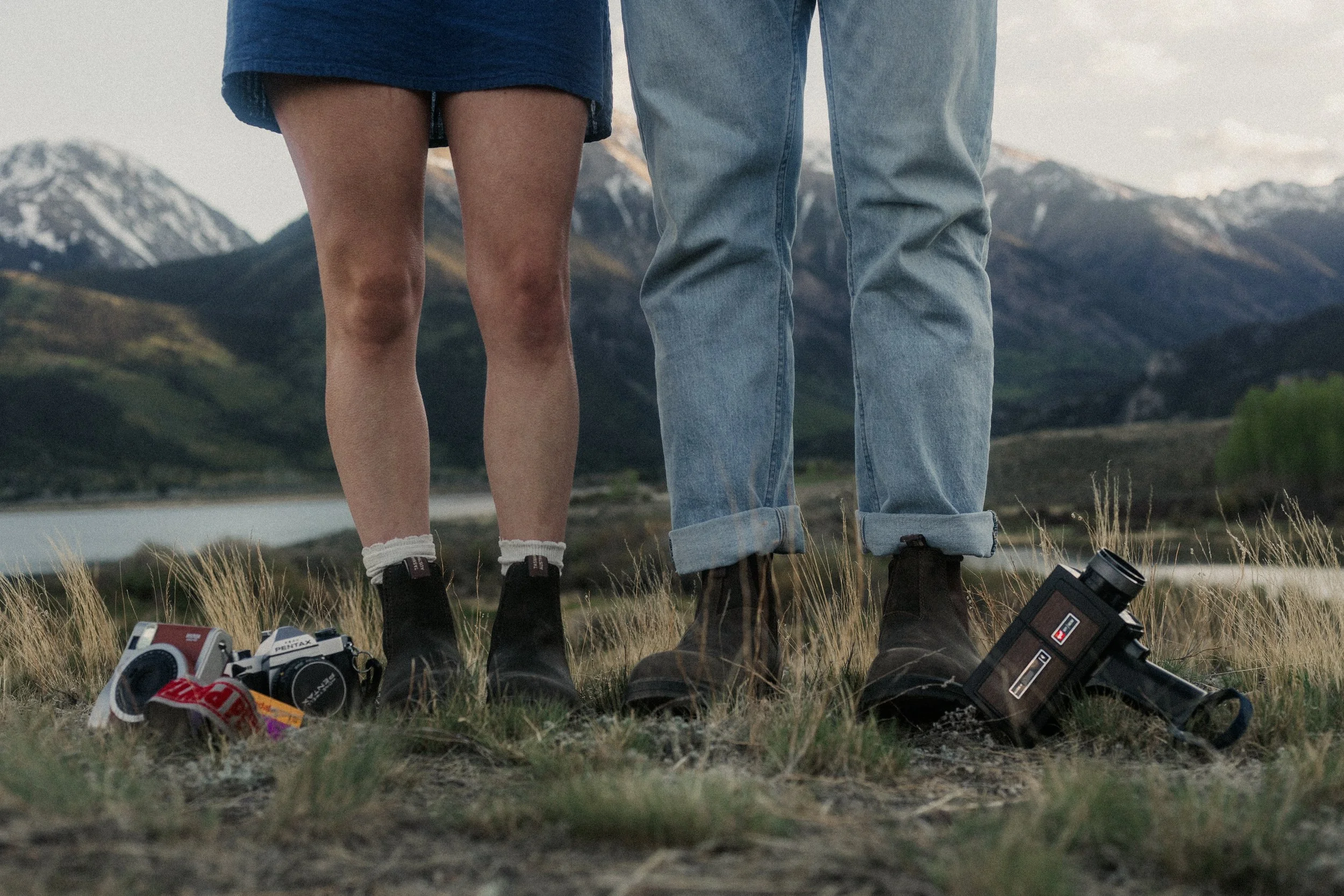 Photo showing the lower bodies of two people standing outdoors on grass with a mountain landscape in the background. One person is wearing a blue skirt and white socks with black shoes, the other is wearing rolled-up blue jeans and brown boots. Sever