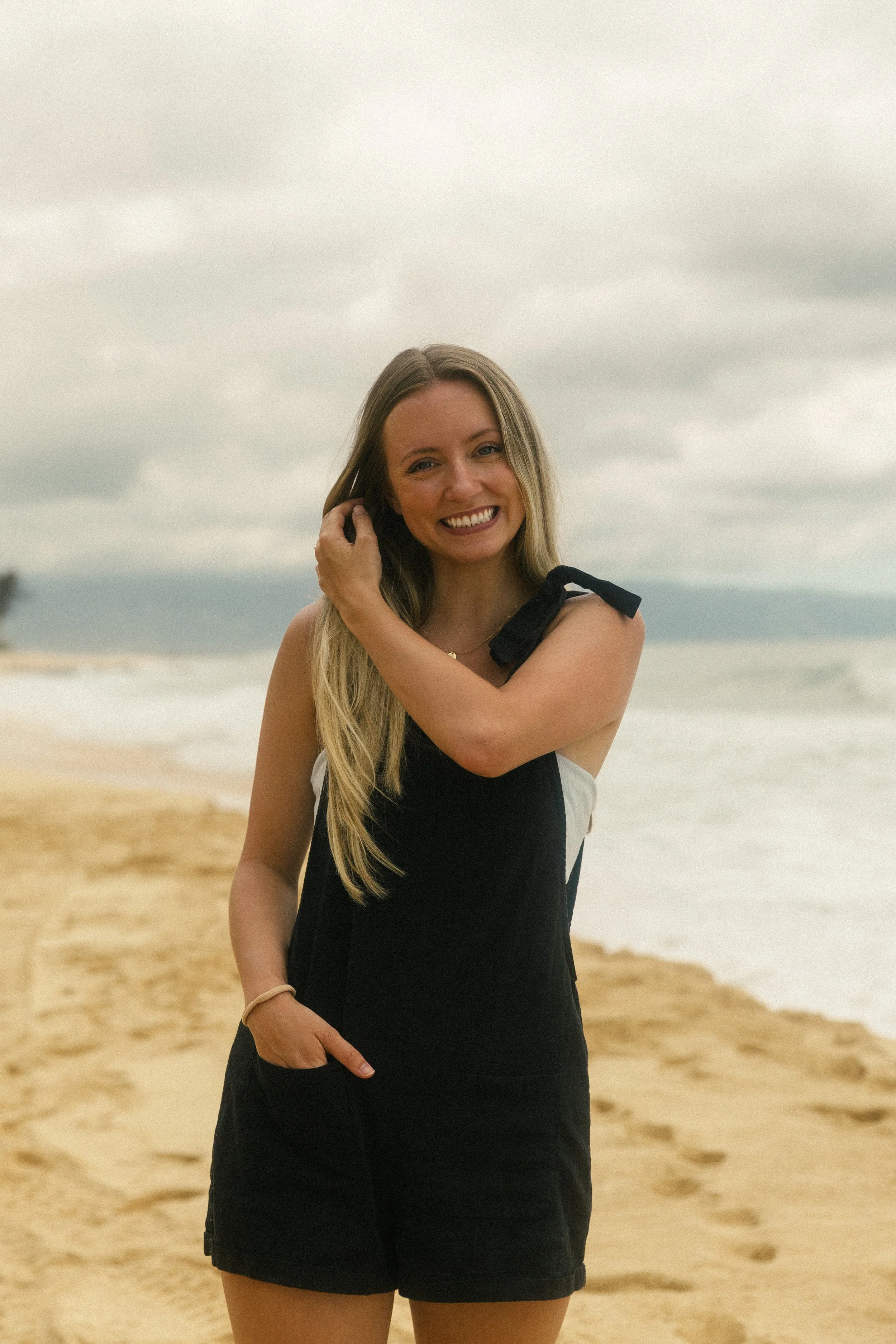 A woman with long blonde hair smiling on a sandy beach with cloudy sky in background, wearing a black and white dress.